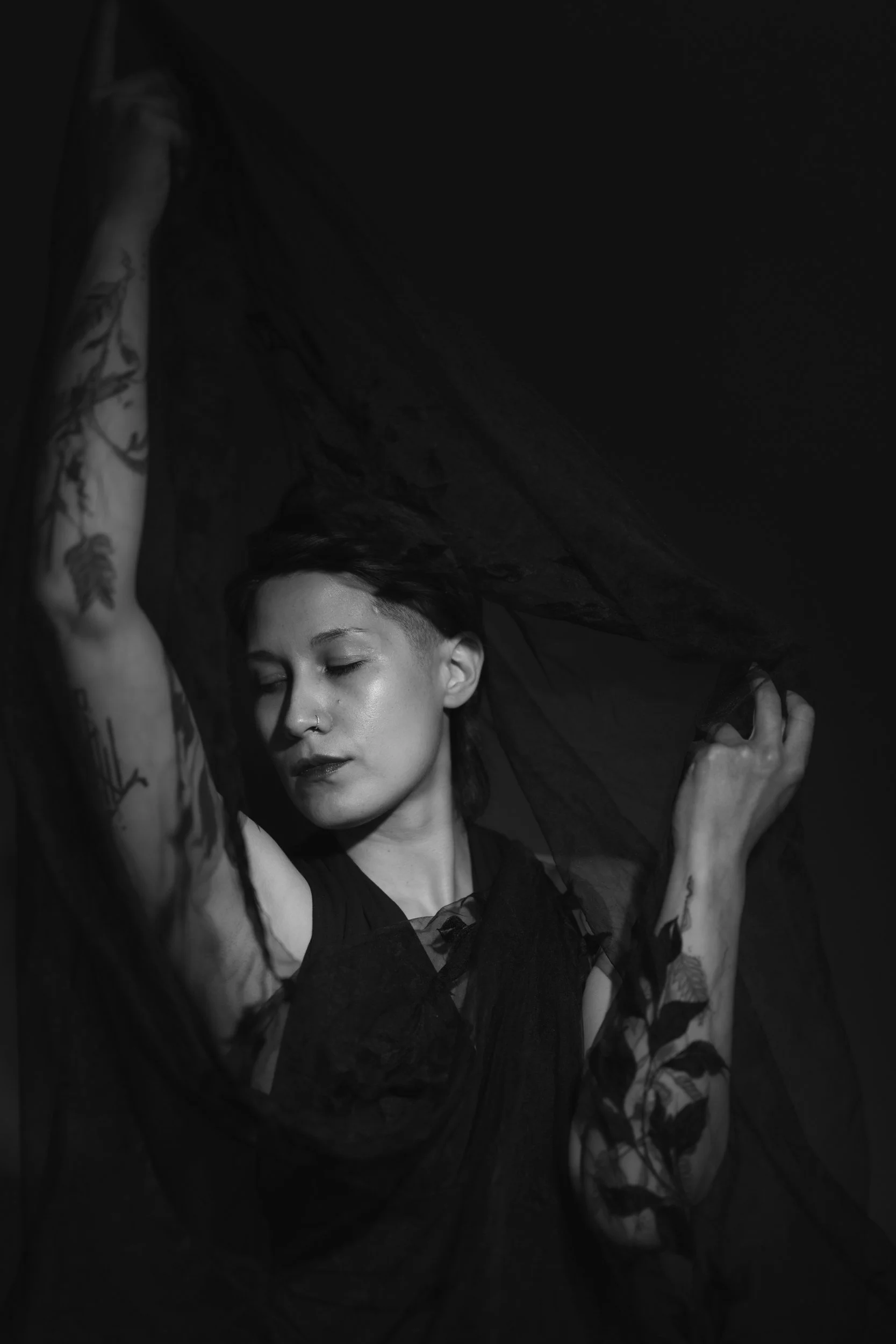 A black and white photo of a person with short hair and tattoos, holding fabric or clothing above their head with their eyes closed.