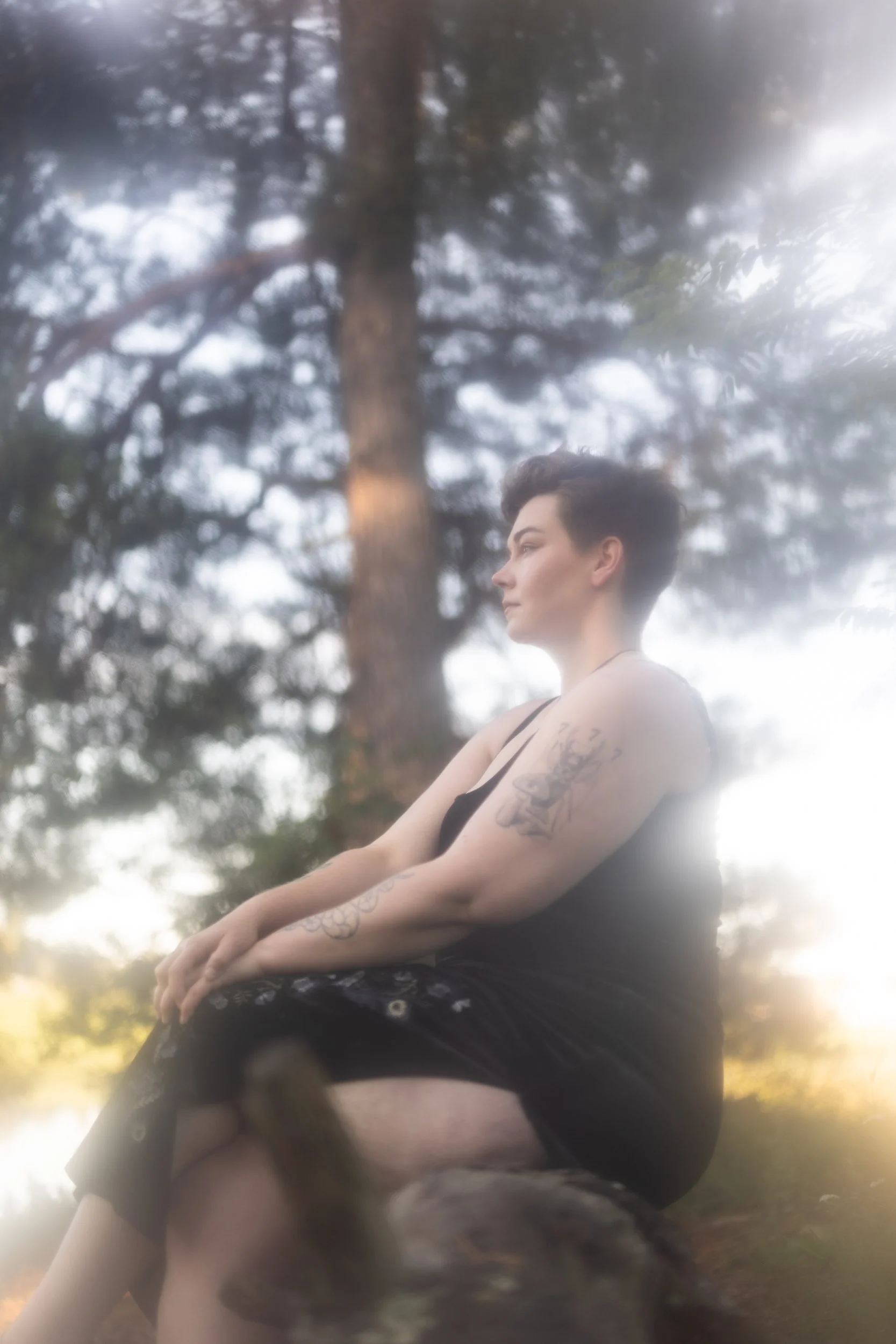A person with short dark hair and tattoos on their arms sitting outdoors with tall trees in the background, captured during sunset or late afternoon.