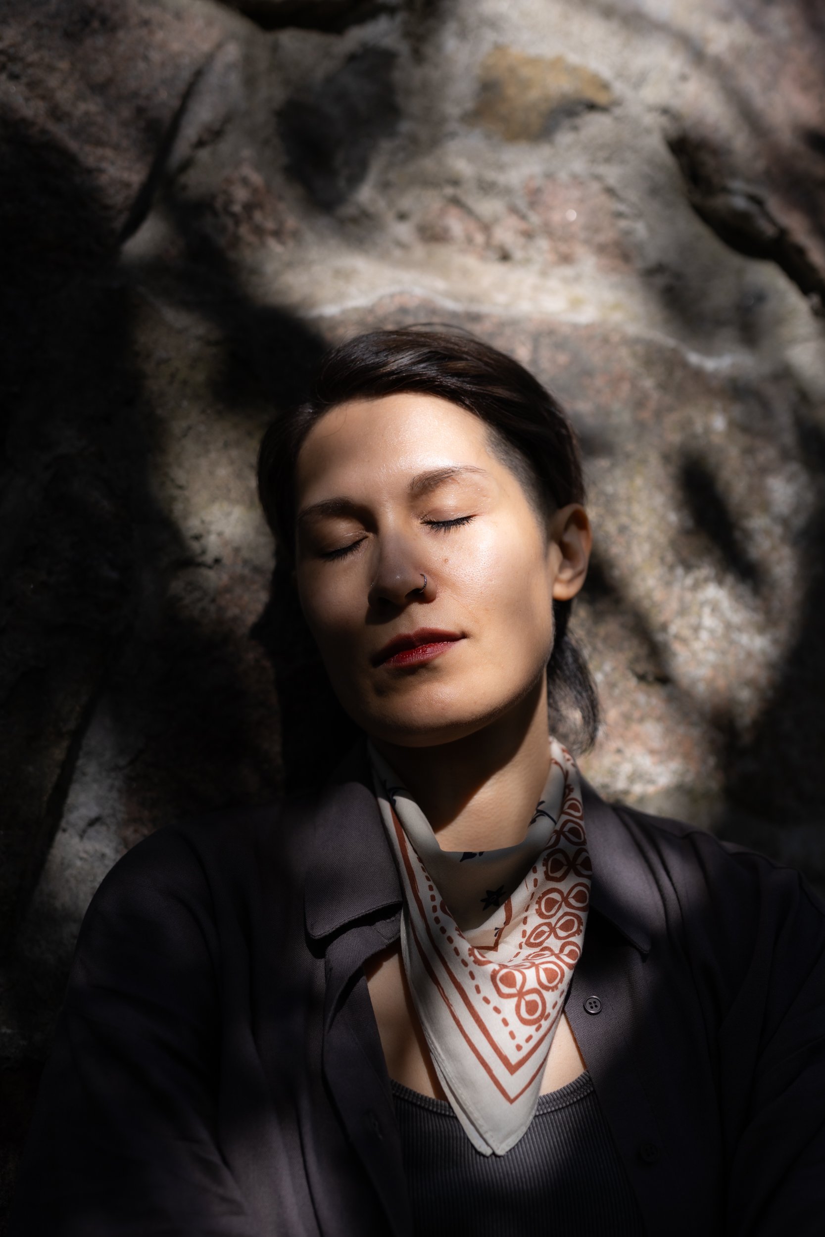A person with short dark hair and a nose ring is resting against a rocky surface with her eyes closed, wearing a black jacket, a patterned scarf, and a black top.