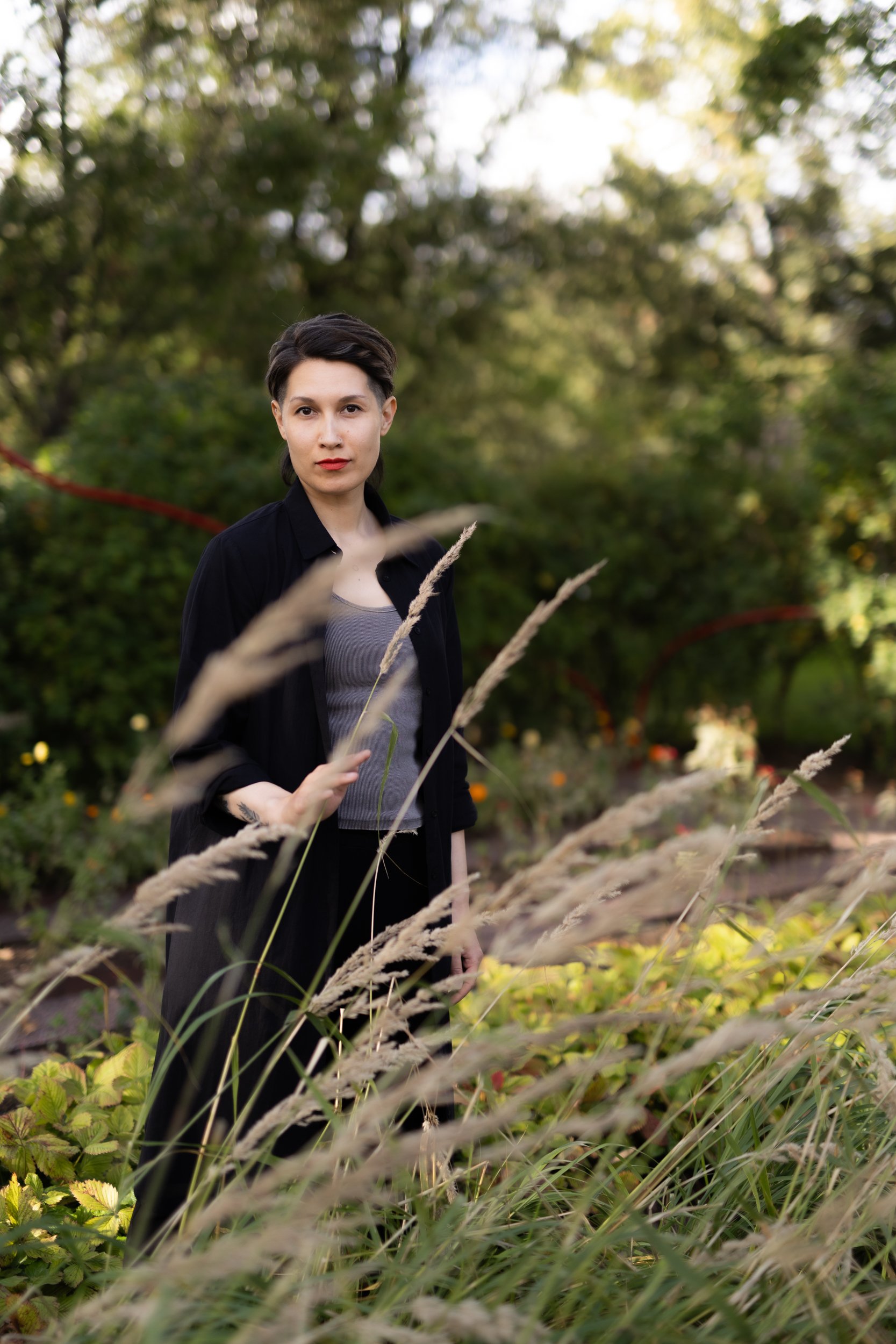 A person with short dark hair, wearing a black blazer and gray top, stands in a lush garden with green plants and trees, holding some tall grasses.
