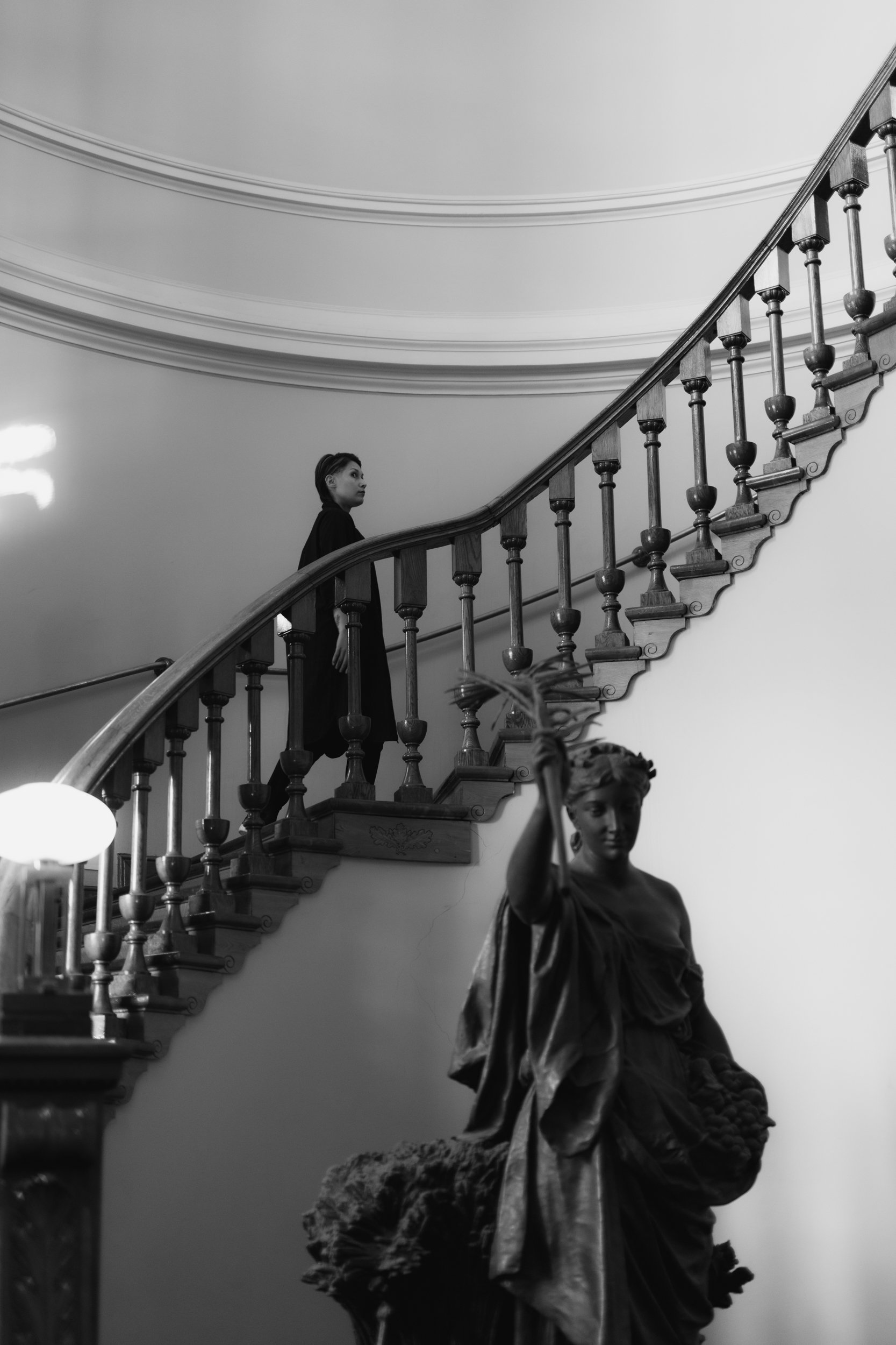 A statue of a woman dressed in a flowing gown stands at the bottom of a curved staircase with wooden balustrades, while a person  in dark clothing stands on the stairs facing away. The scene appears indoors with ornate ceiling molding.