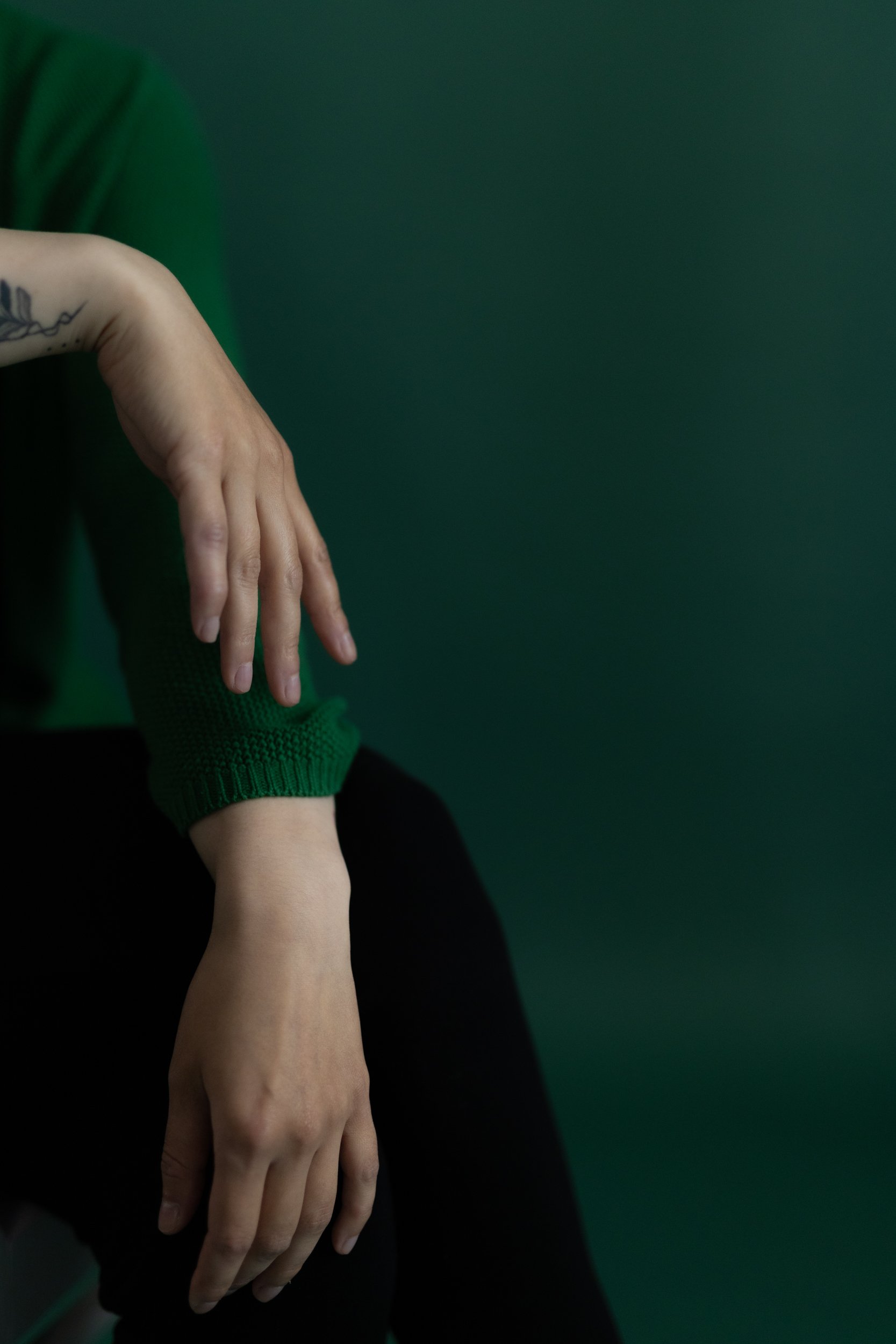 Close-up of two clasped hands with tattoos, against a dark green background. One hand is resting on the other's wrist.