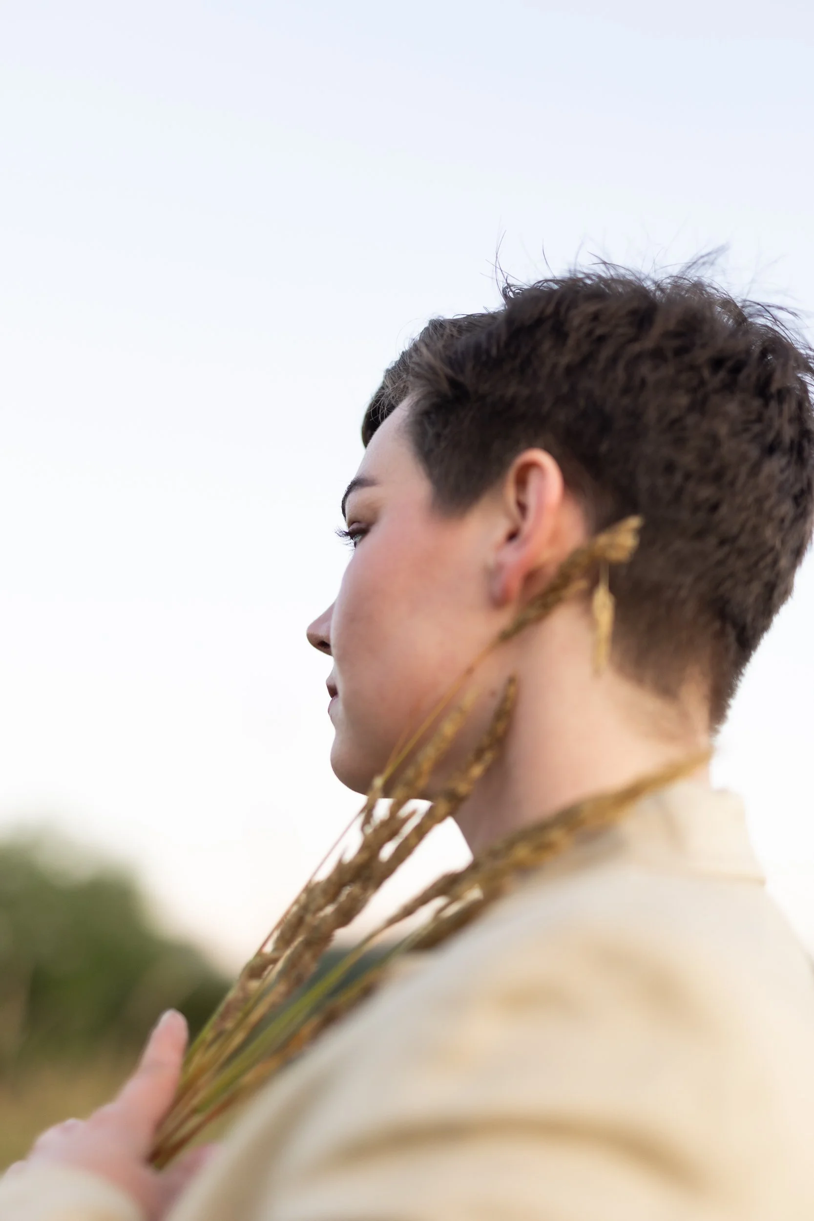 A person with short dark hair in profile holding dried wheat stalks, with a soft outdoor background and a pale sky.