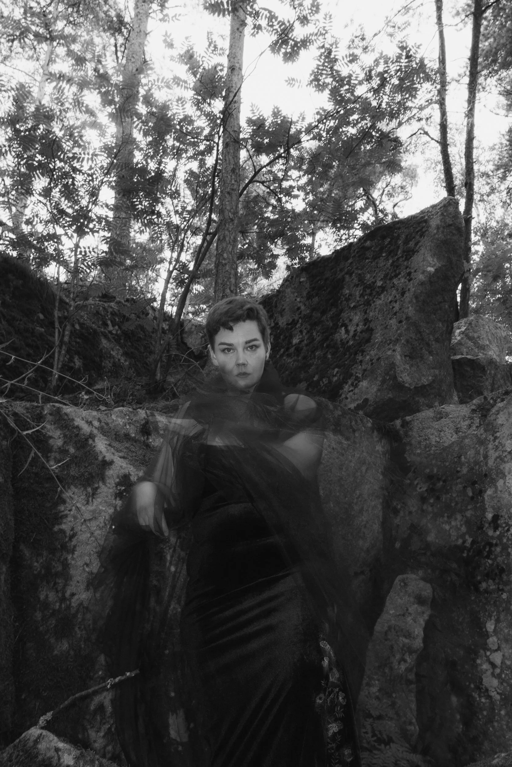 A black and white photo of a person with short hair sitting among rocks and trees in a forested area.
