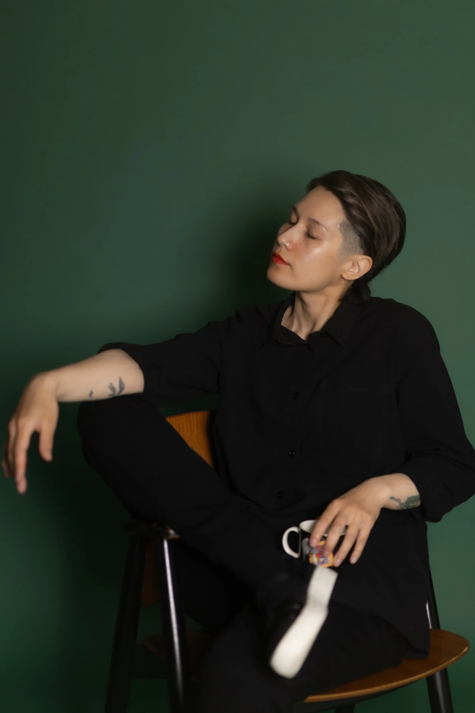 A person with short dark hair, wearing a black shirt, sitting in a wooden chair against a dark green wall. Their eyes are closed, and they have a tattoo on their left arm. They are holding a coffee mug in their right hand.