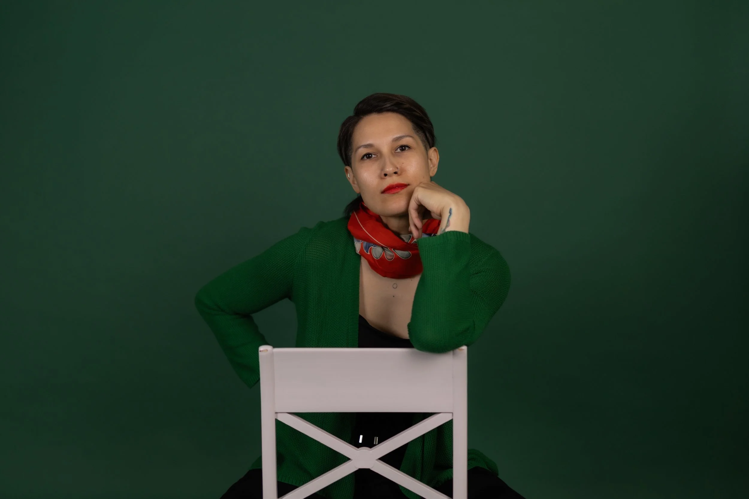 A person with short dark hair and red lipstick sitting behind a white chair against a solid green background. They a resting her face on the left hand, with the elbow on the table, wearing a green cardigan, a red patterned scarf, and a nose piercing.