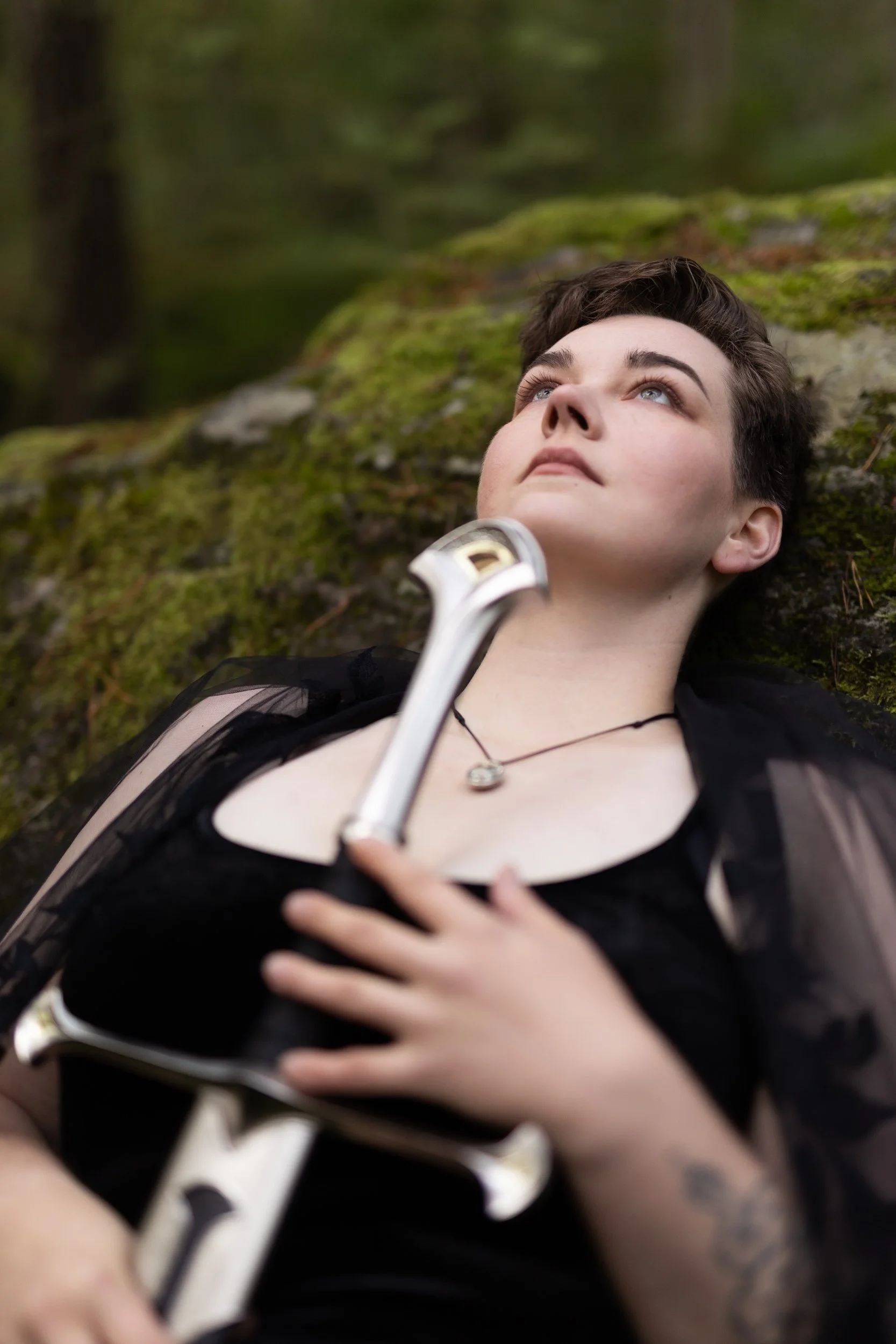 Person lying on mossy ground surrounded by trees, holding a wrench on their chest, wearing a black outfit and a necklace, with short dark hair, in a forest setting.