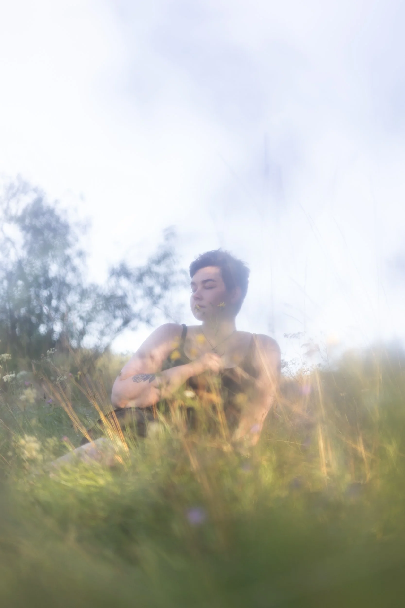 A person with short dark hair and tattoos on their arm, standing outdoors among tall grass, with a cloudy sky and trees in the background. They are wearing a black tank top and have yellow flowers on their face.