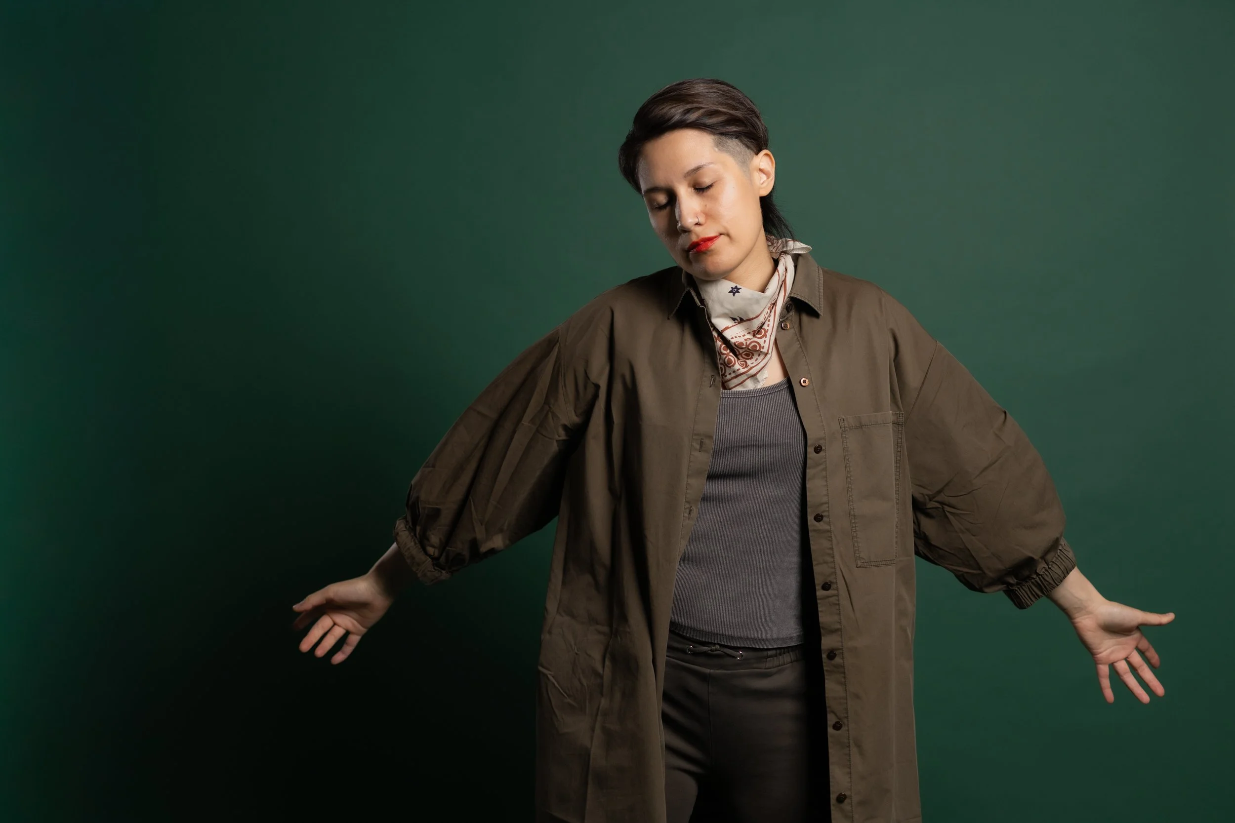 A person with short dark hair, wearing a brown jacket over a gray top and a white bandana, standing against a green background with arms slightly raised and eyes closed.