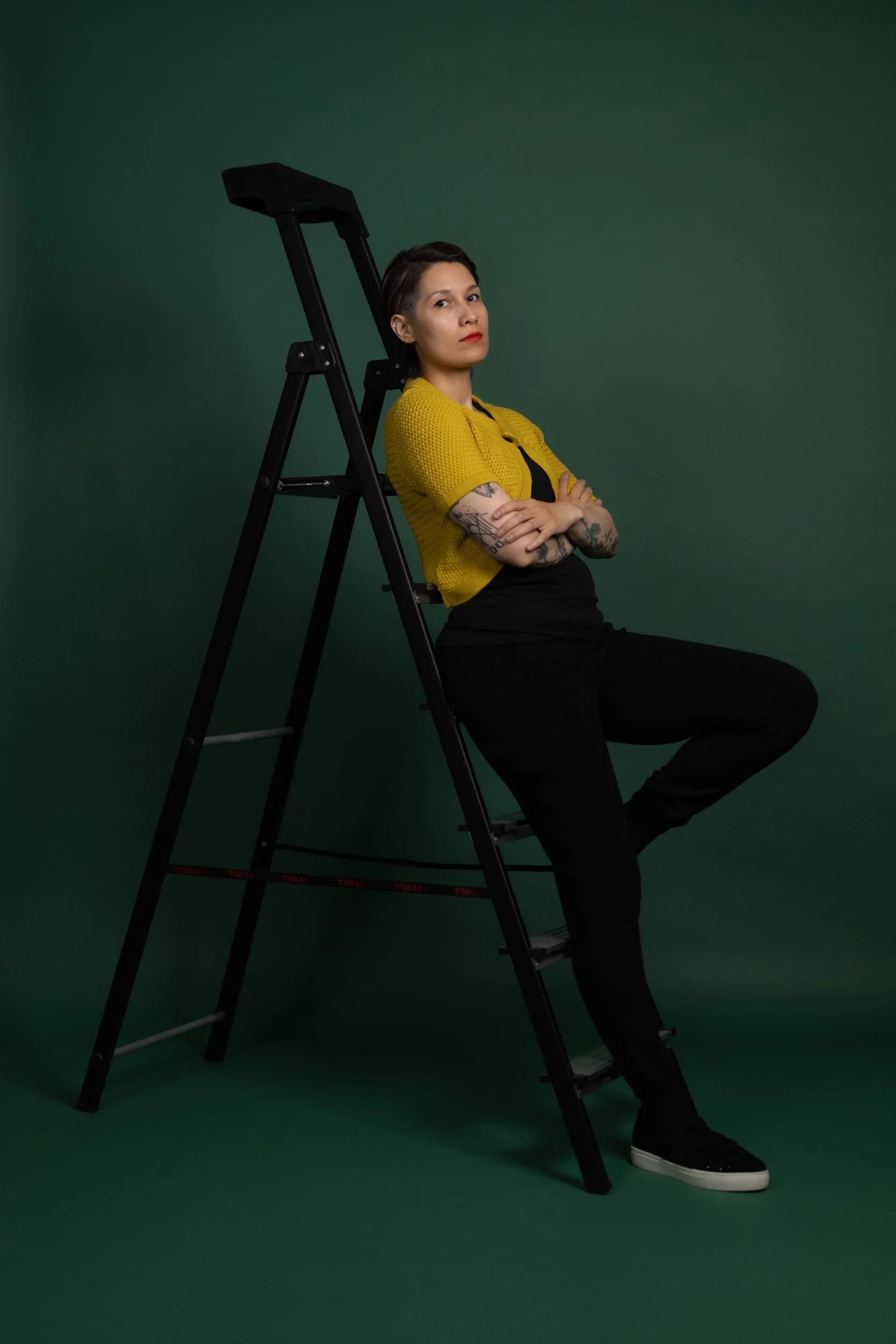 A person with tattoos wearing a yellow sweater and black pants, sitting on a stool with crossed arms, leaning against a black ladder in front of a green backdrop.
