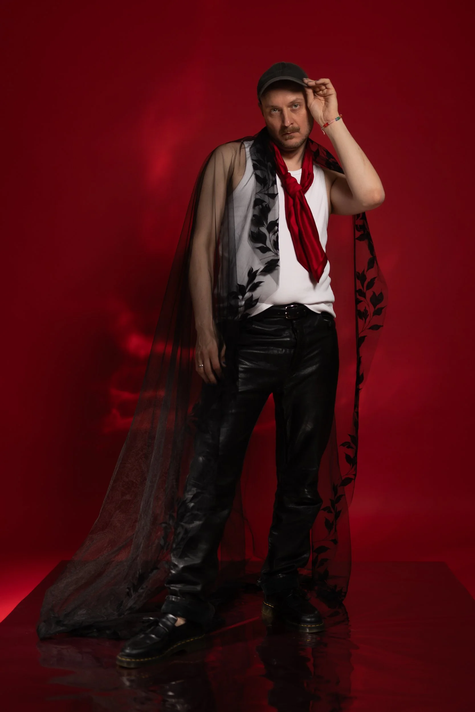 A strong, self-aware portrait in which the person stands in front of a red background. A black veil rests on their shoulders, and they hold one hand to their temple. The pose is contrapposto. The person is wearing black leather pants and a white slee