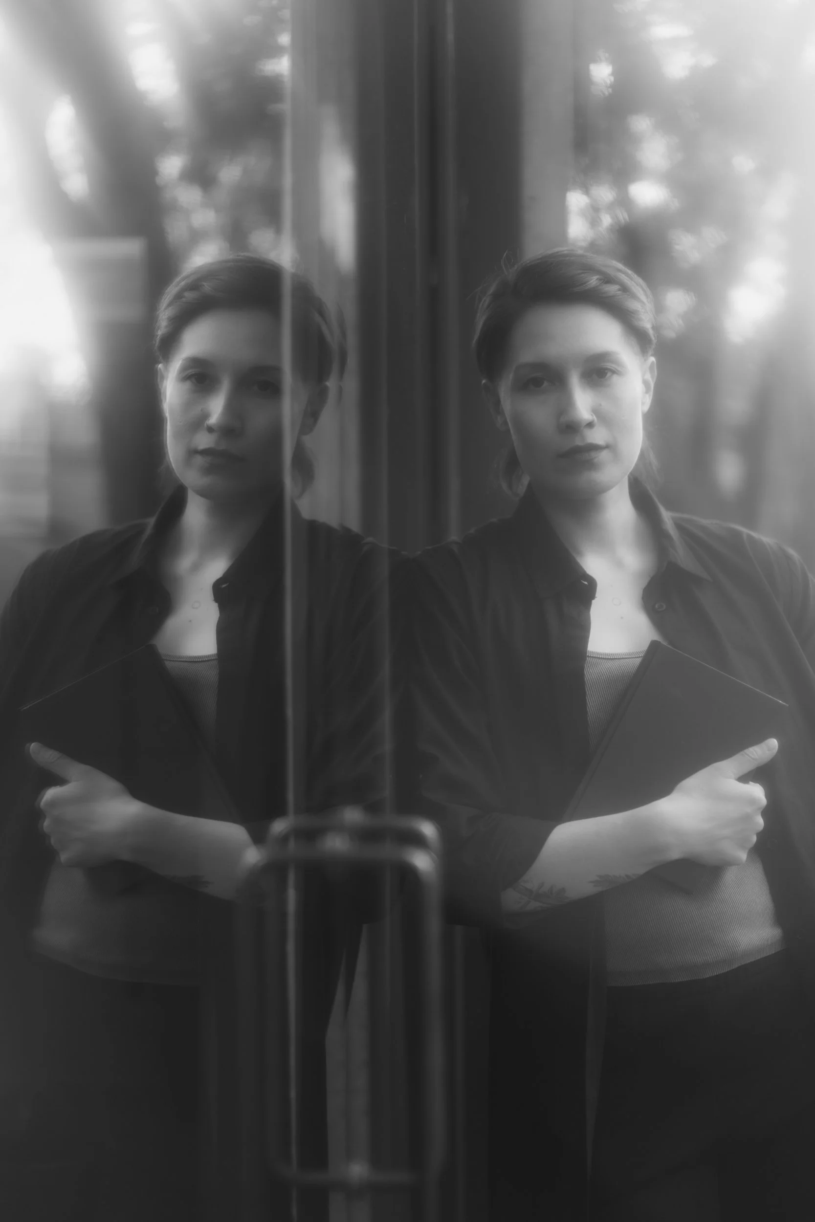 Black and white photo of a person taking a selfie in front of a mirror, with her reflection visible. They have short hair, are wearing a dark shirt, and is holding a notebook with her arms crossed.
