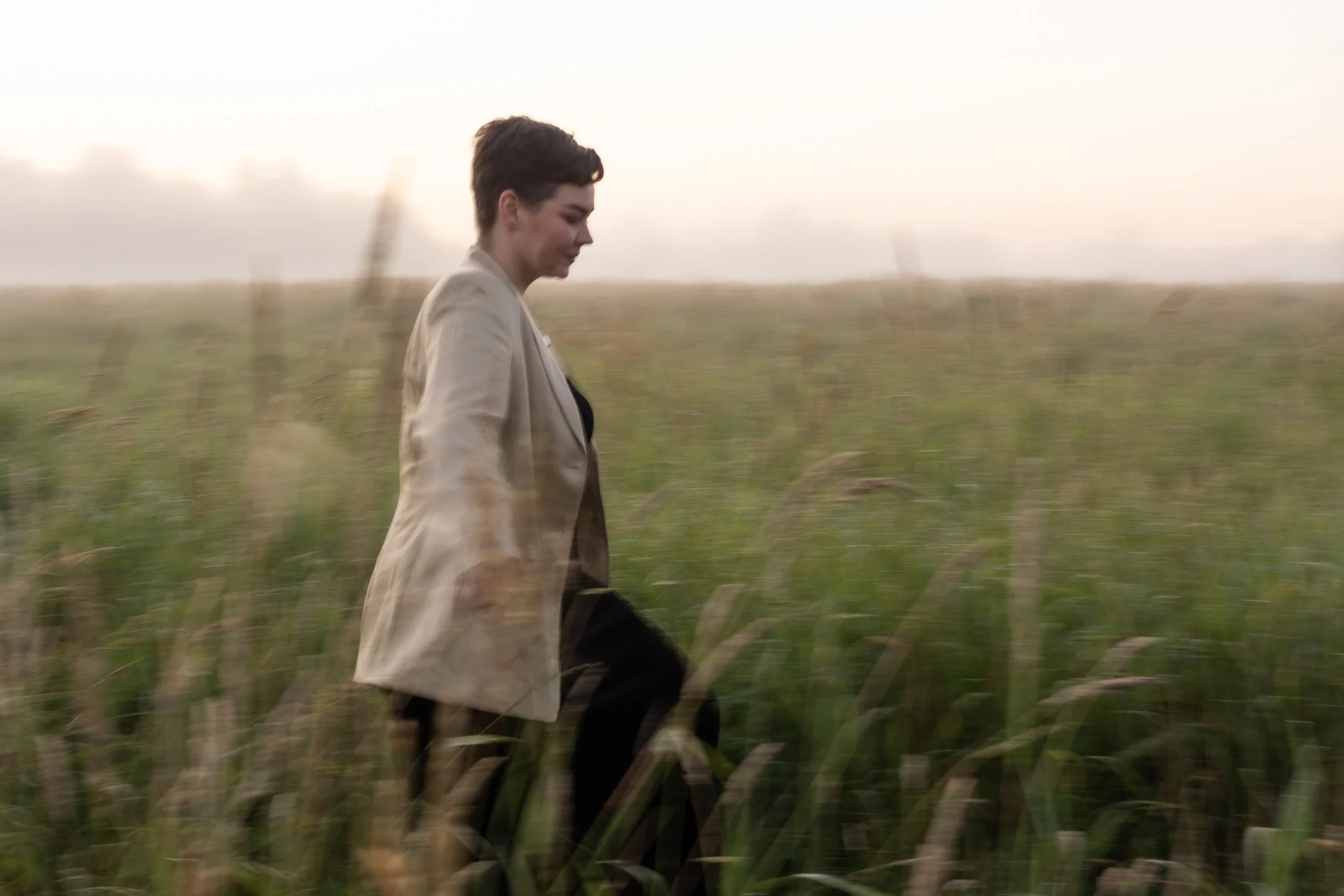 Person walking through a grassy field at sunset, wearing a beige blazer and black pants.