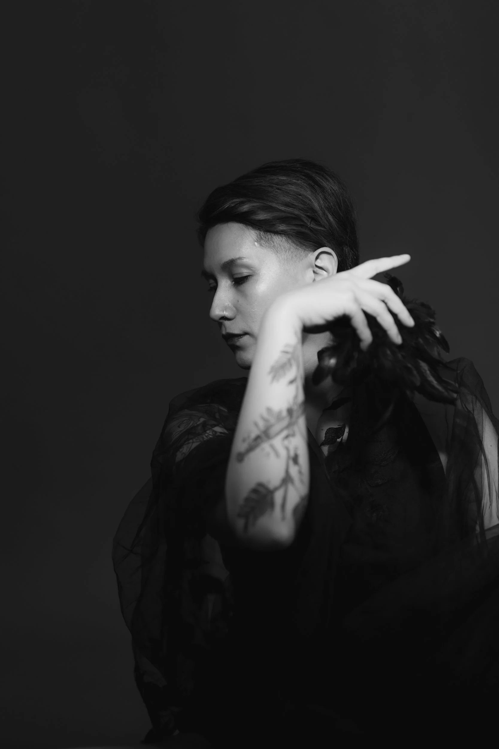 A black and white photo of a person with short hair and tattoos on their arm, posing with their hand behind their head against a dark background.