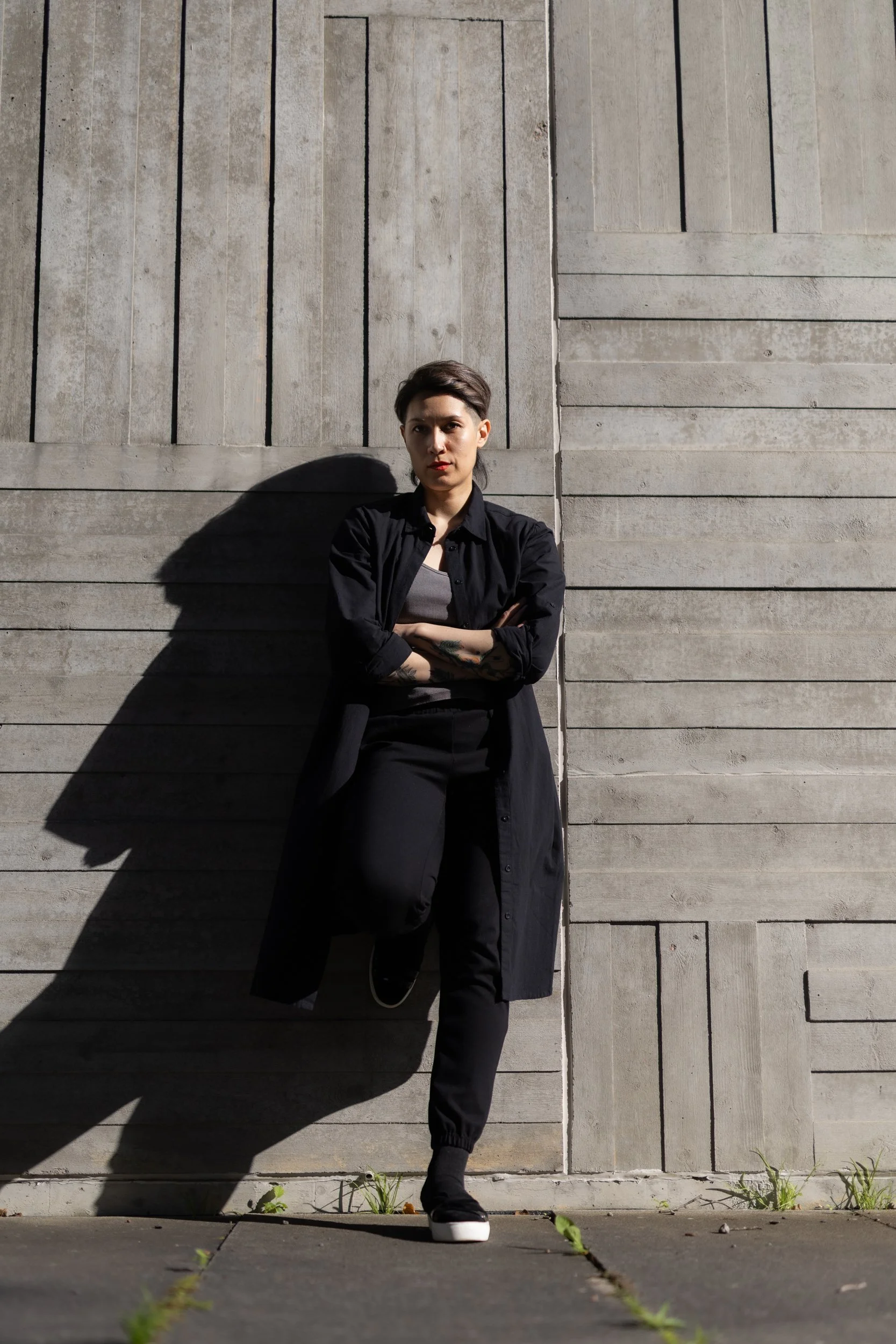 A person with short dark hair, wearing a black jacket over a gray top, black pants, and sneakers, standing against a concrete wall with crossed arms and looking at the camera. Sunlight casts a shadow on the wall behind.
