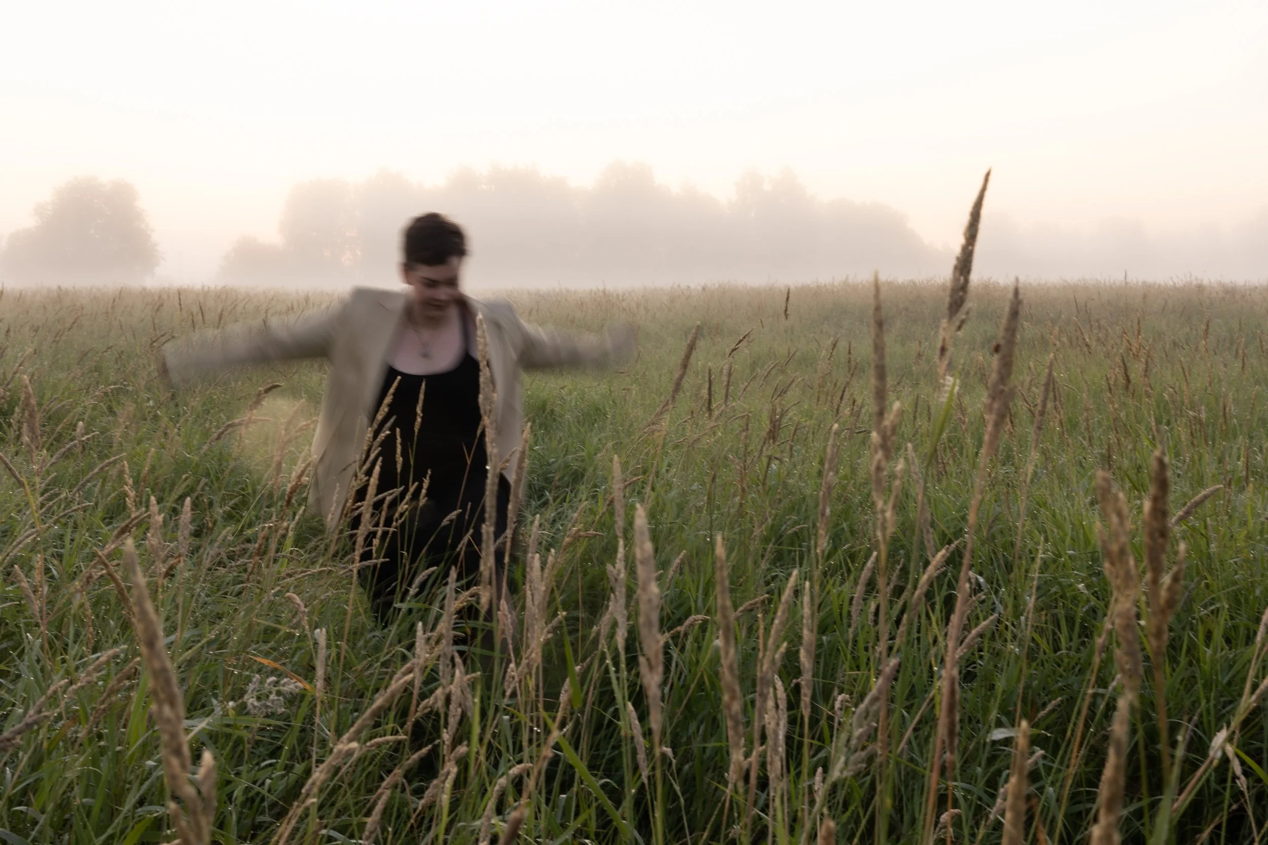 A person walking through a grassy field with tall grass and trees in the background, foggy morning atmosphere.