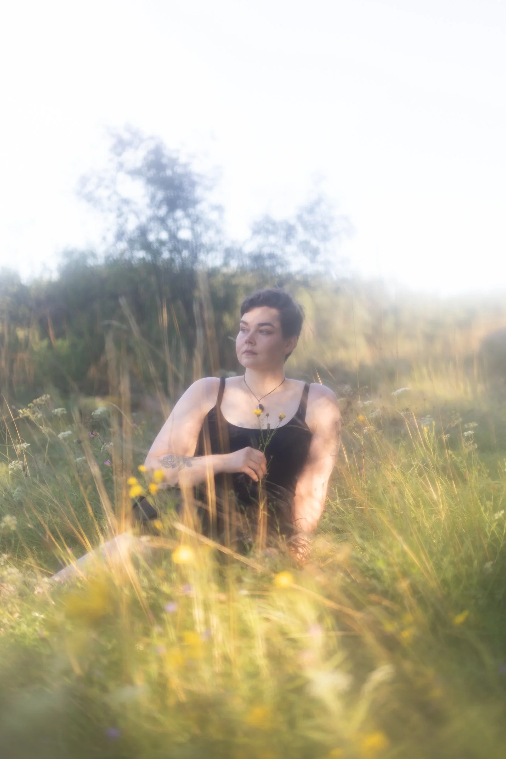 A person with short dark hair and tattoos ot their arm sits in a field of tall grass and wildflowers, holding a small yellow flower and looking to their right with a serene expression, with trees and a bright sky in the background.