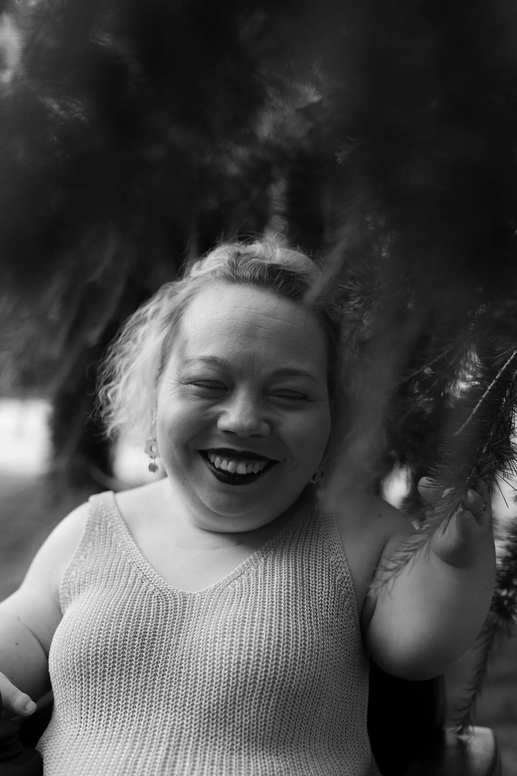 A woman laughs and smiles in the picture. She is frameed by spruce branches and she has a big smile on her face. She has a blond bob haircut and looks very happy. She is short-statures and uses a wheelchair.