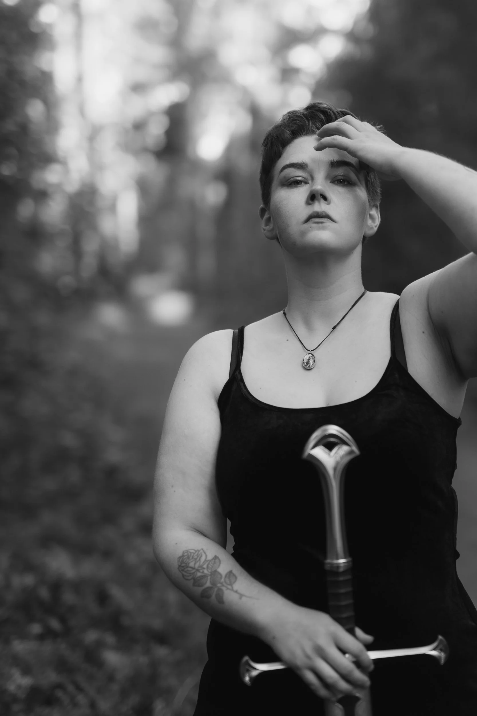 Black and white photo of a person with short hair, wearing a black tank top and a necklace, standing outdoors with trees in the background. They have a tattoo on their left arm and are holding a sword.