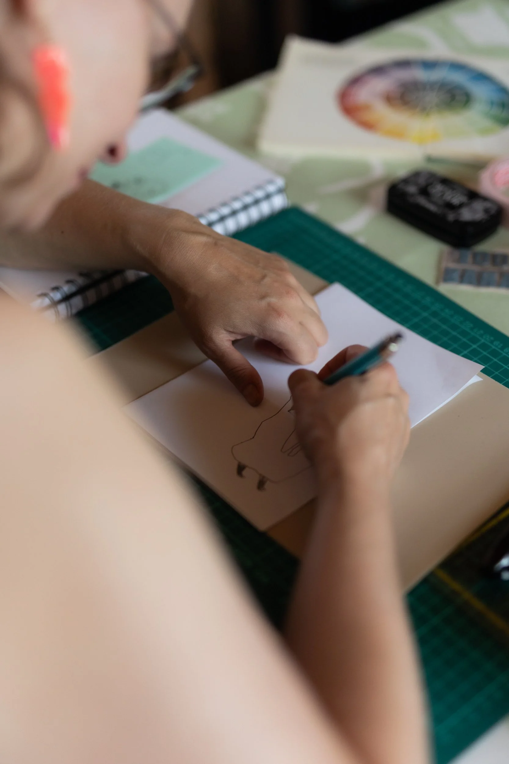A person is drawing on a piece of paper at a desk surrounded by coloring and art supplies, including a color wheel and various supplies.