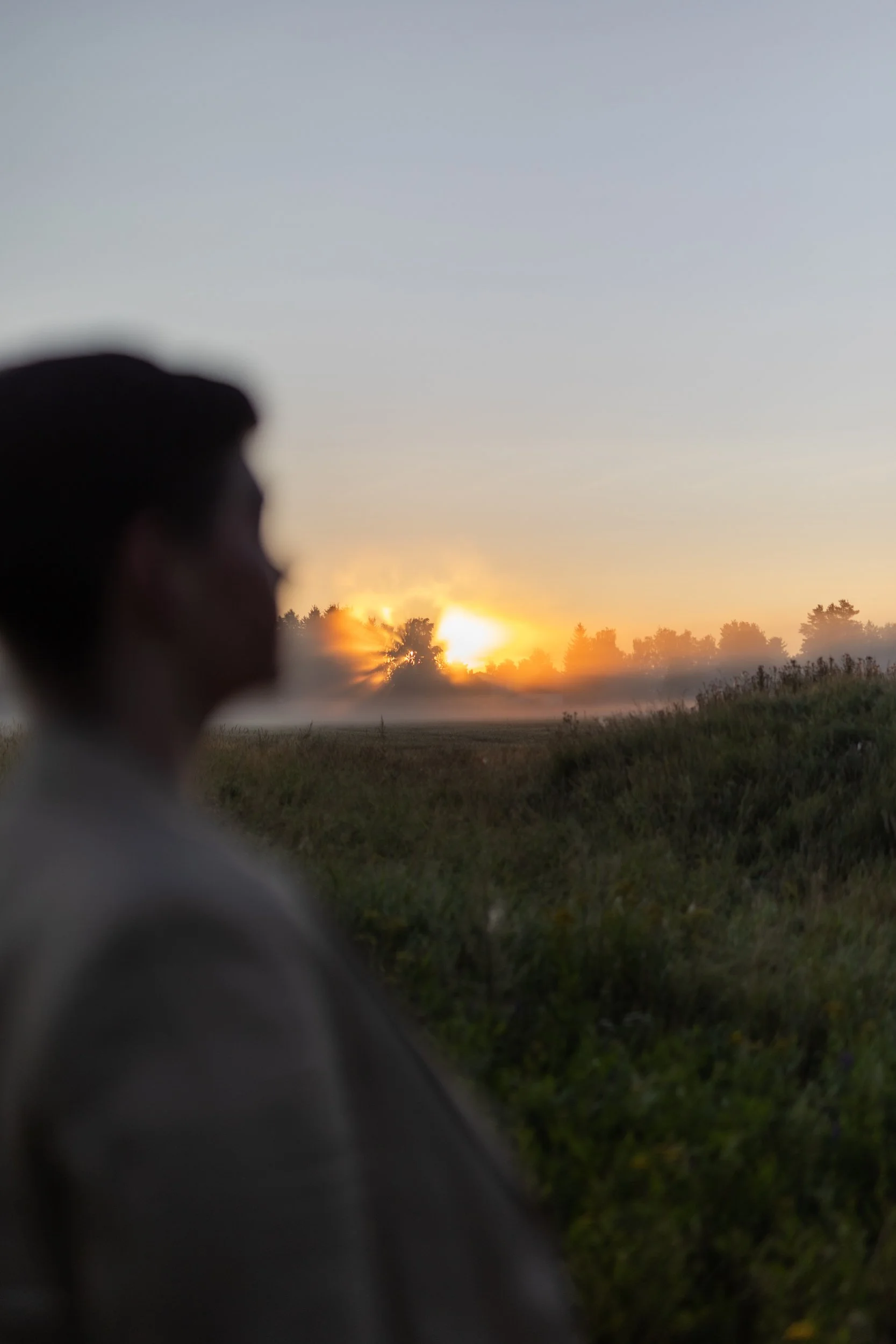 A person sitting outdoors at sunset or sunrise, with trees and open field in the background, and a dog visible in the foreground.