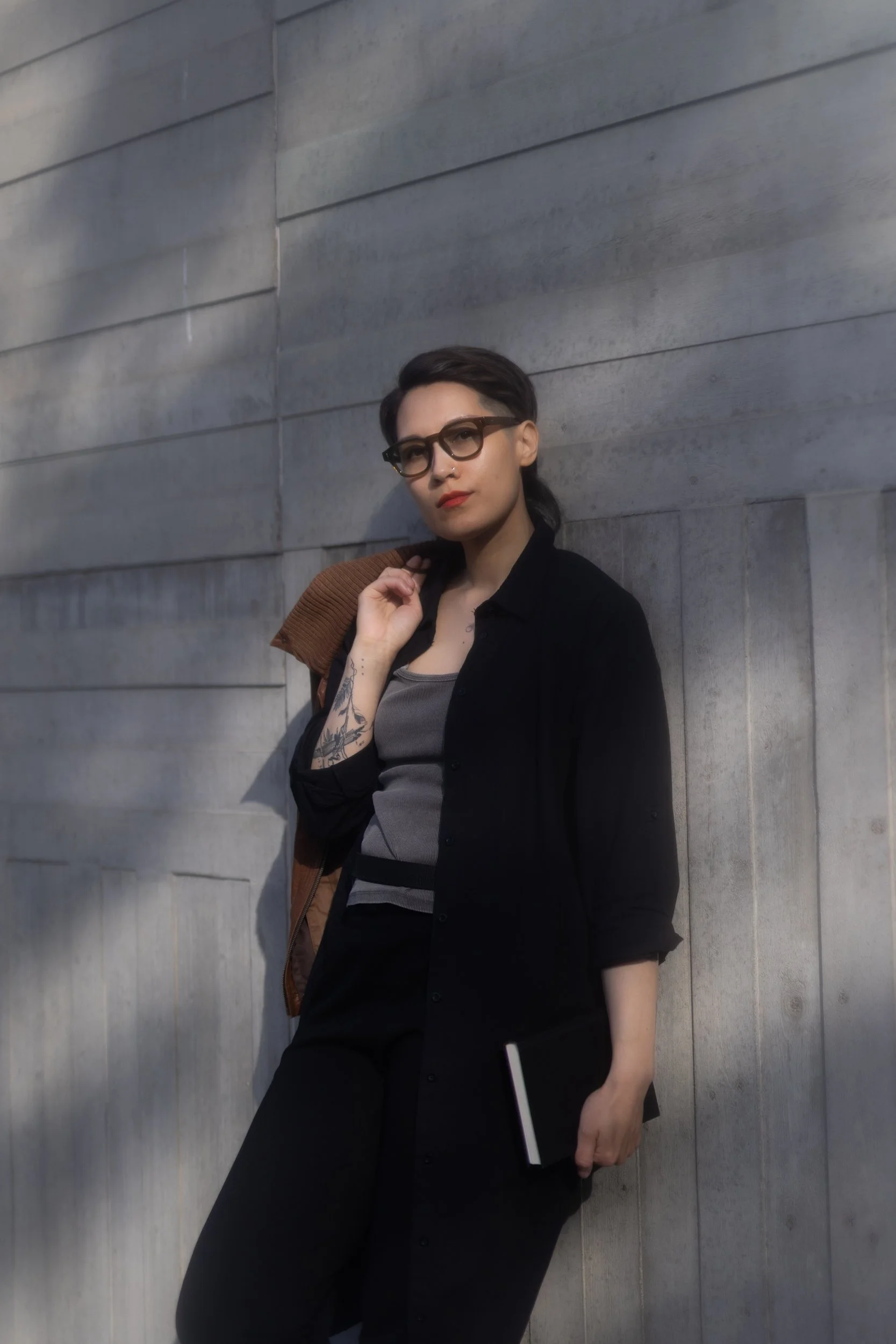 A person with dark hair, glasses, and tattoos on their arm, dressed in a black shirt and gray tank top, standing against a gray tiled wall. They are holding a book or notebook in her right hand and has a brown jacket draped on their shoulder.