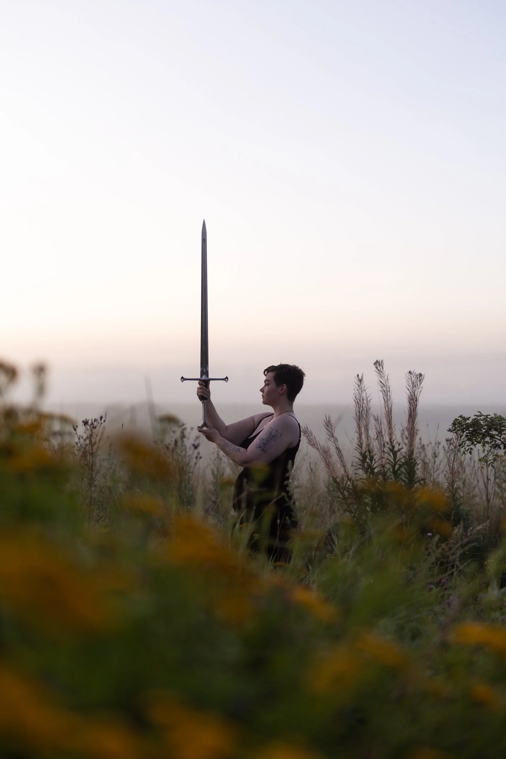 A person with tattoos on their arm stands in a field of tall grass and plants, holding a sword upright with both hands, during sunset or sunrise with a clear sky.