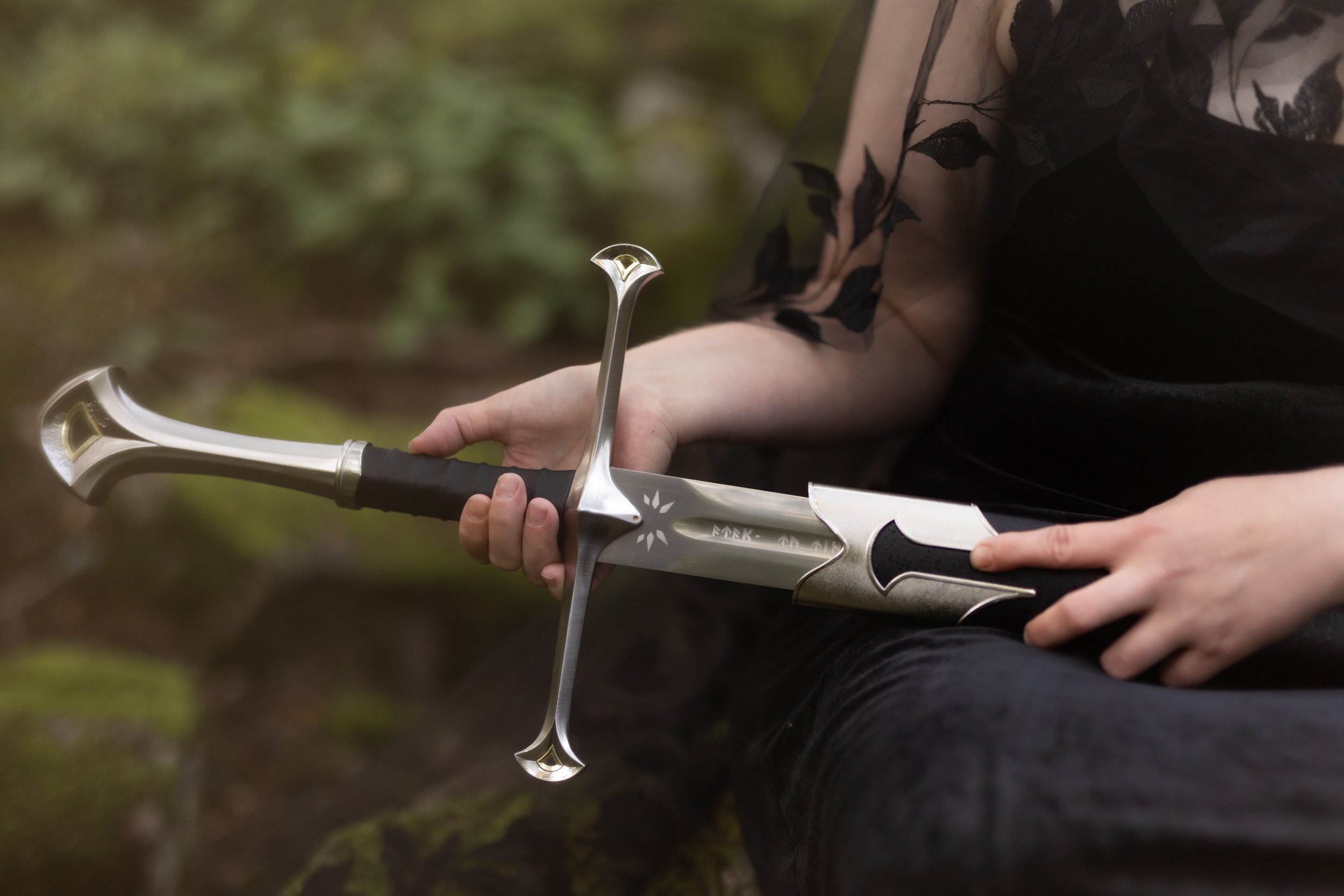 A person dressed in black holding a decorative sword with a silver hilt and blade, standing outdoors with blurred greenery in the background.