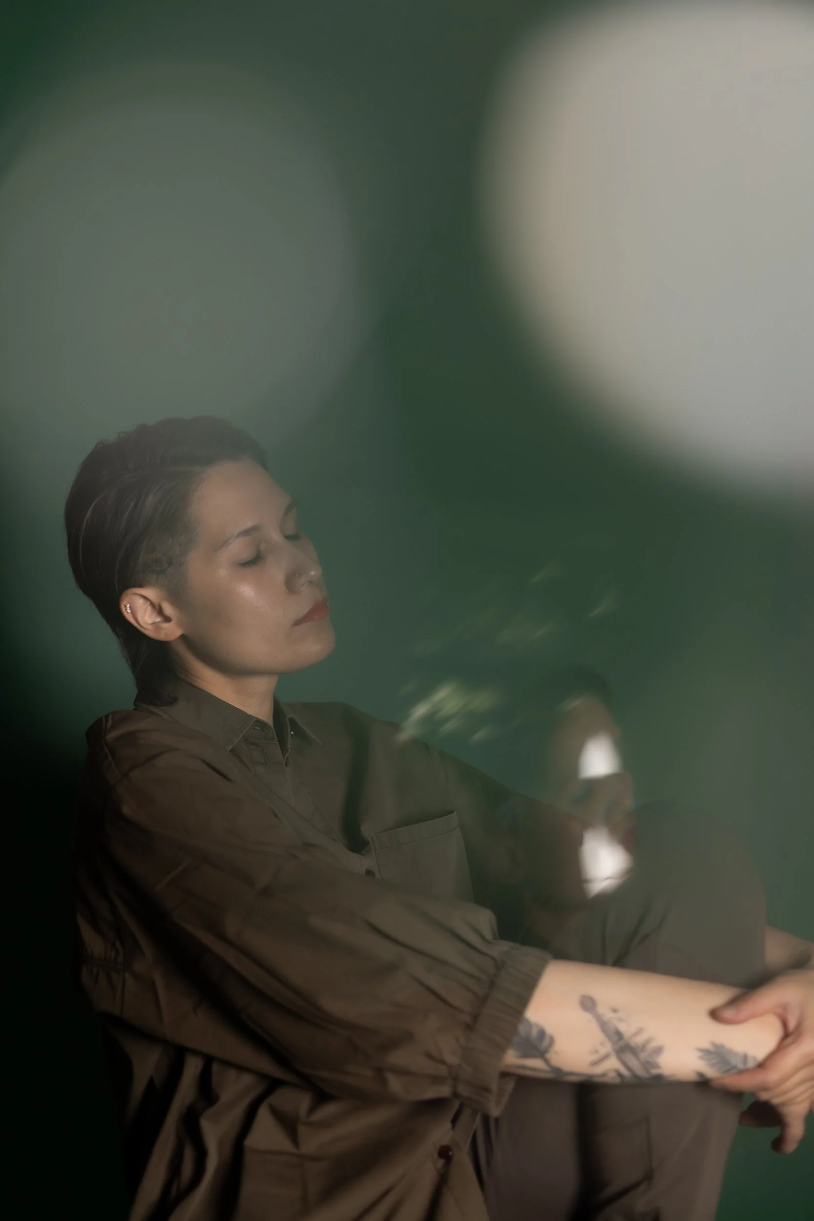 A person with short dark hair, closed eyes, and a nose piercing, sitting with their arms crossed, wearing a brown shirt, in front of a green background, with a tattoo of leaves on their left forearm.