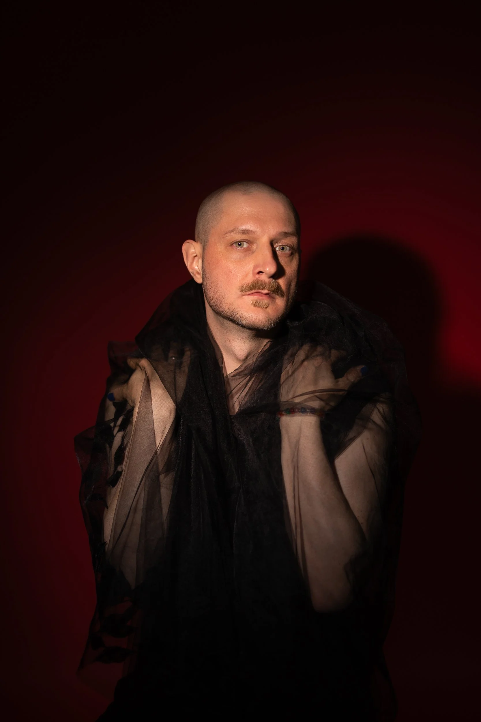 A dramatic and emotionally evocative image in which Mikko Toiviainen is dressed in a black mesh shirt and has a black veil on his shoulders. He holds his hands on his shoulders and looks directly into the camera. The lighting is soft but dramatic.