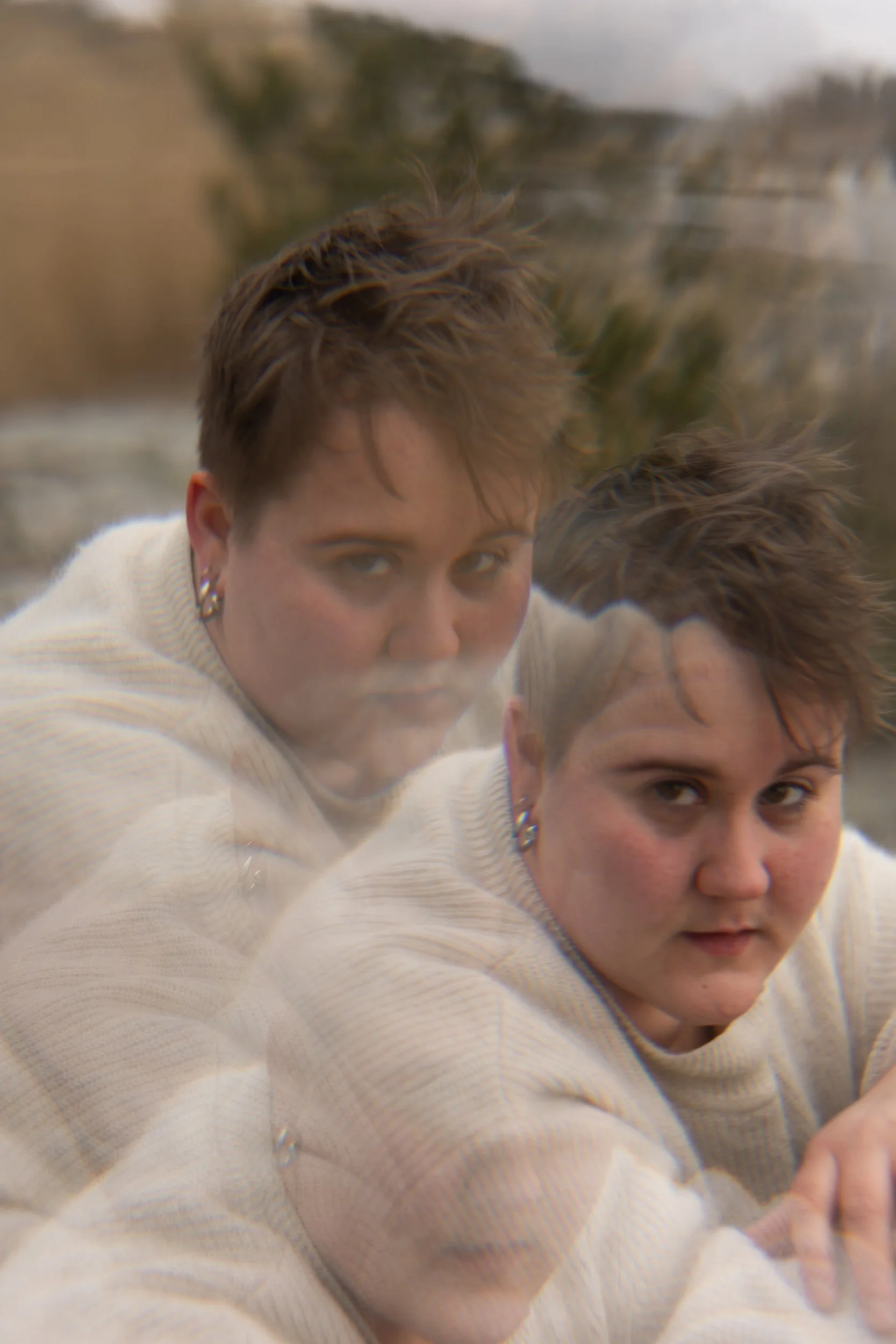 In this intensive portrait a non-binary person looks straight at the camera. Theey have a white shirt on and silver earrings. The image of the person is multipled in three times in a way that they overlap themself in the photo.