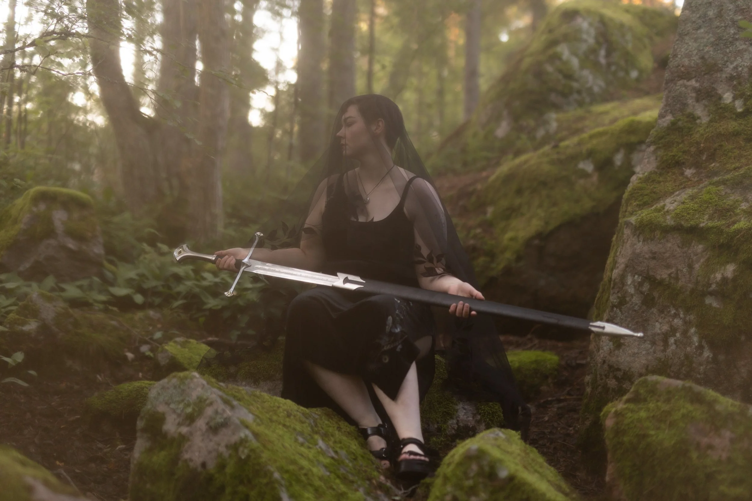 A person dressed in black with long dark hair and a black veil sits in a mossy forest holding a sword. The scene is mystical with filtered sunlight through the trees.