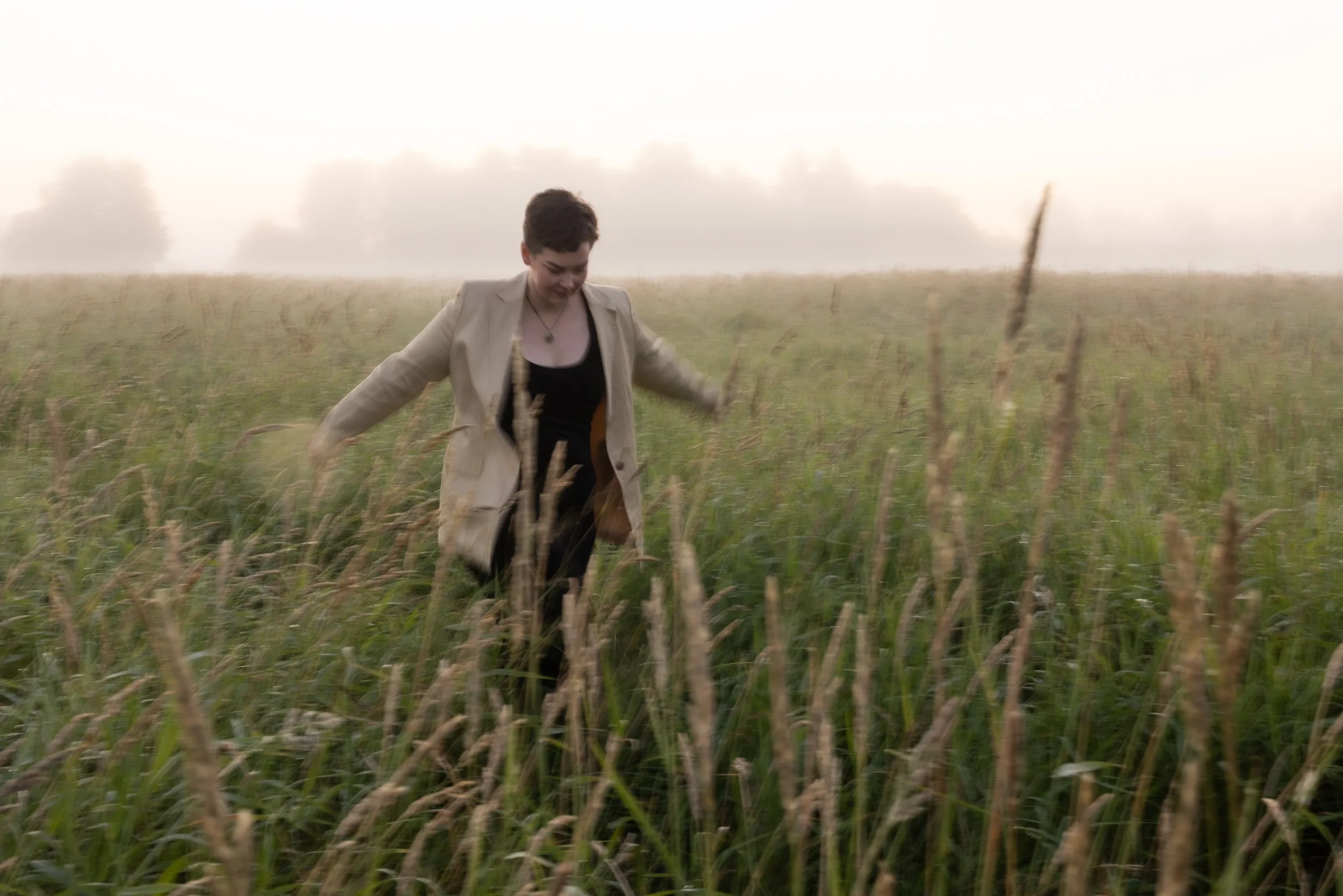 A person with short hair in a beige jacket and black outfit walking through a grassy field during foggy weather.