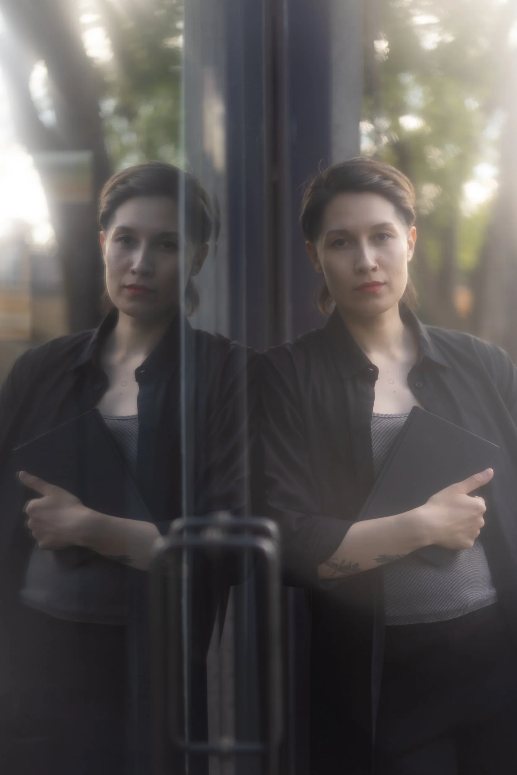 A person with short hair standing in front of a window, wearing a black shirt and holding a notebook,  with trees visible outside through the window.