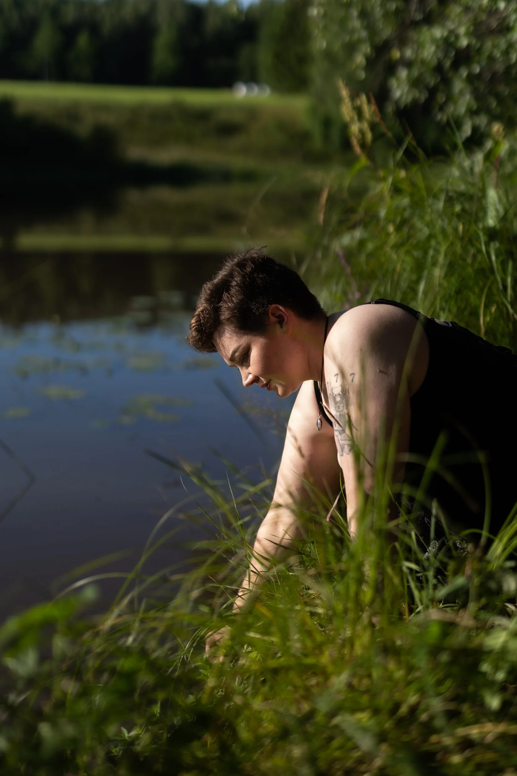 A young person with short brown hair leaning over the edge of a body of water surrounded by green grass and trees in the background, with a blurred side view.