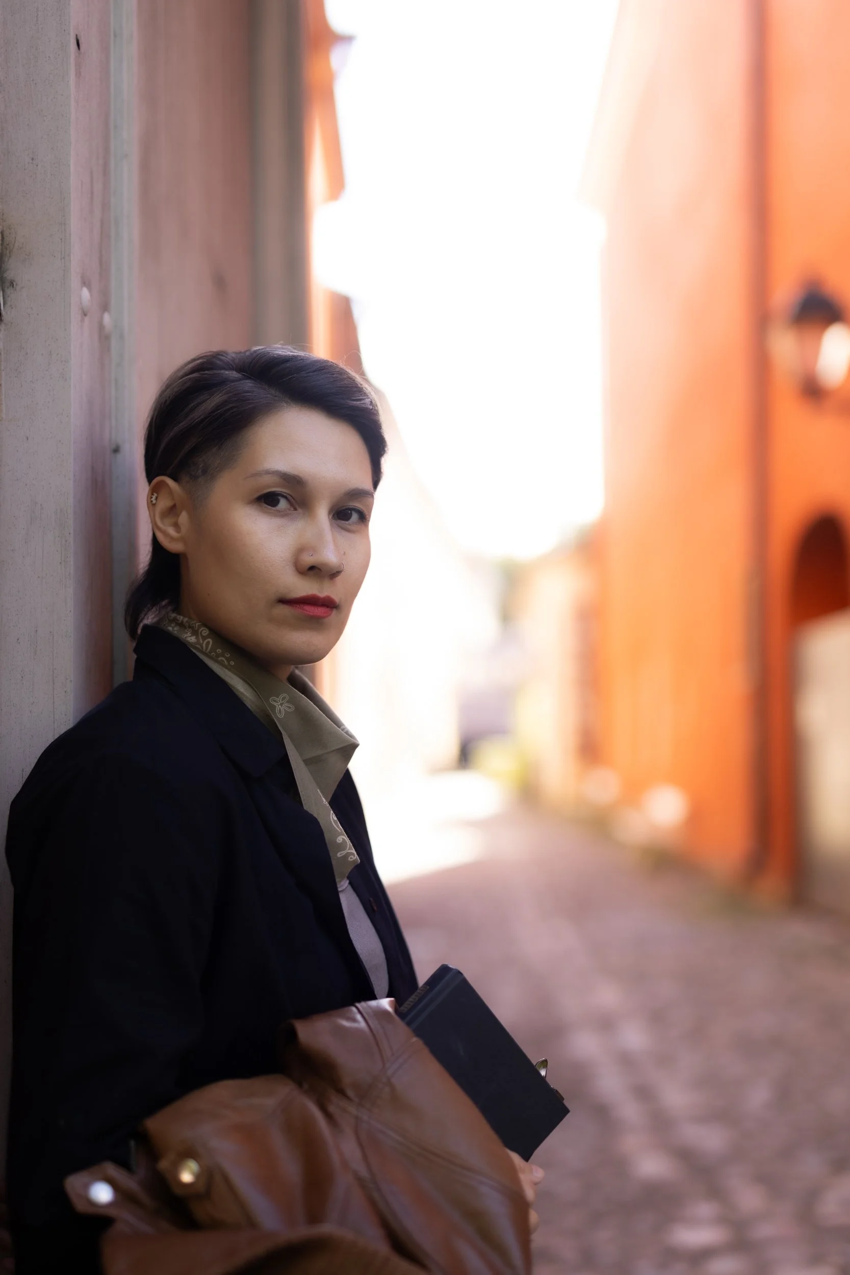 A person with short dark hair  is standing outdoors next to a wooden wall with a bright, sunlit background. They are wearing a black blazer over a green shirt with a patterned collar, holding a black notebook, and have a brown leather jacket.