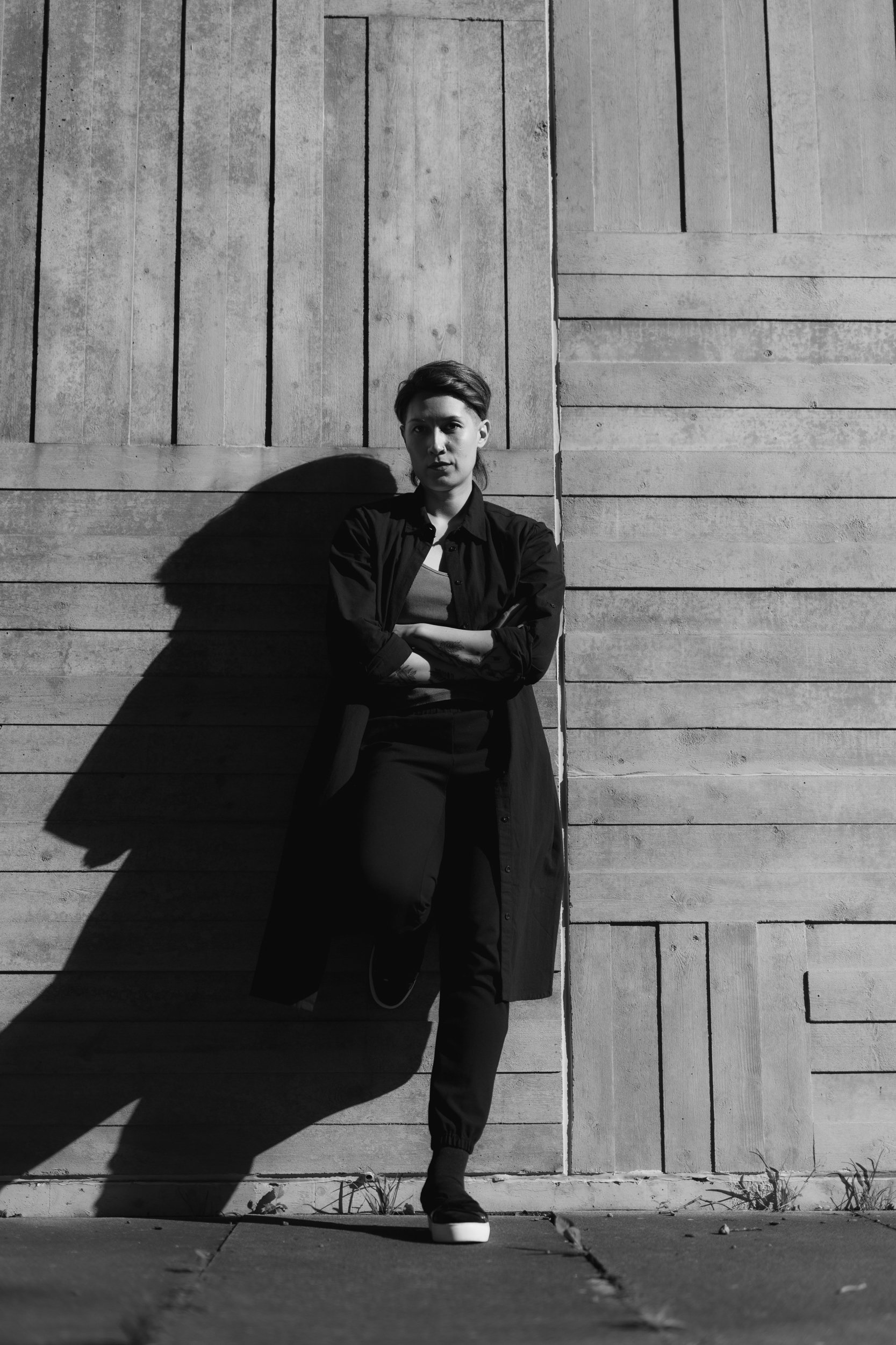 A person with short hair and casual clothing stands with their arms crossed in front of a wooden wall, casting a shadow in black and white.