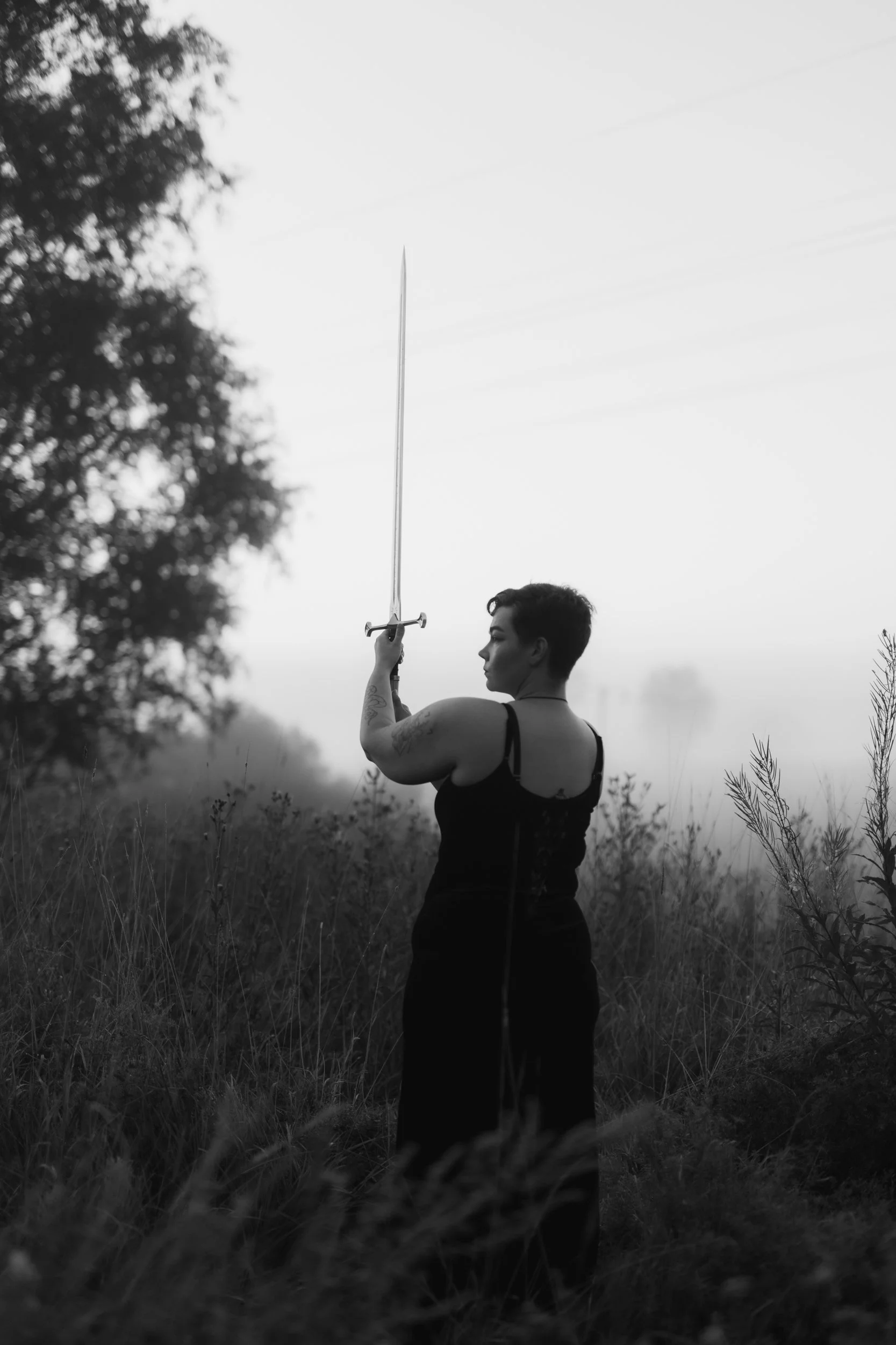 A person with short dark hair and tattoos on their arm, standing outdoors in a grassy area, holding a sword vertically in one hand, with trees and power lines visible in the background, in black and white.
