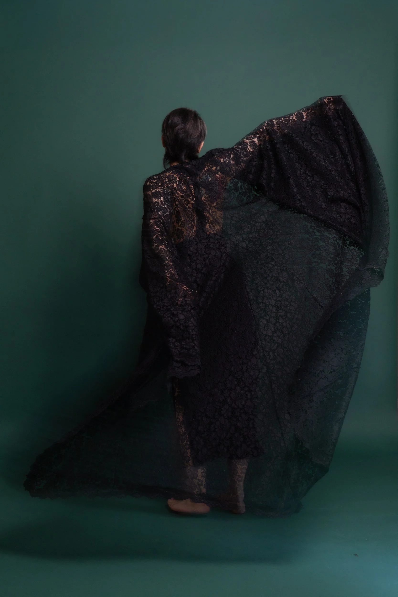 A person with short dark hair wearing a long black lace dress, standing against a green wall.