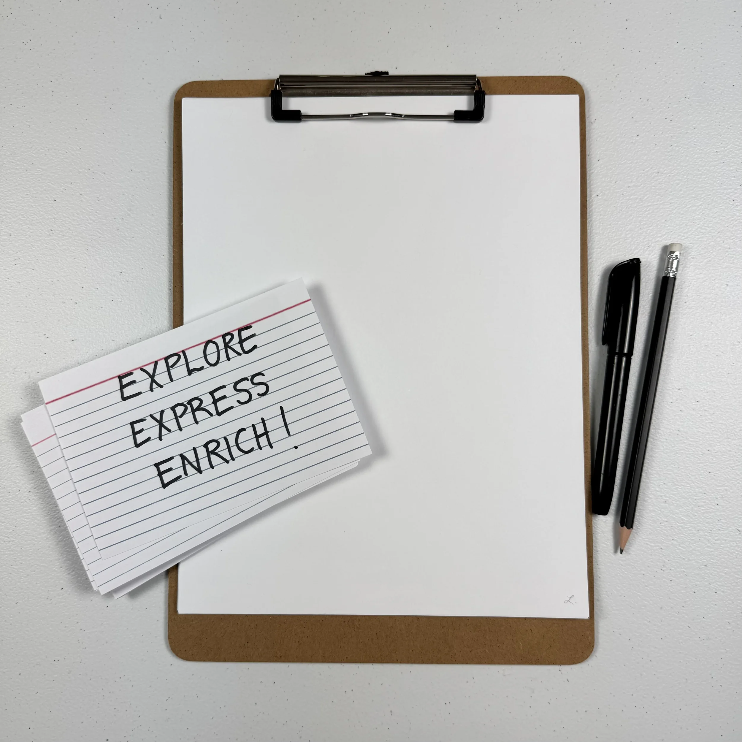 A cork message board with a metal clip holding a blank sheet of white paper, two black pens and a crayon on the right side, and a lined piece of paper with handwritten words that say 'EXPLORE, EXPRESS, ENRICH' in black marker.
