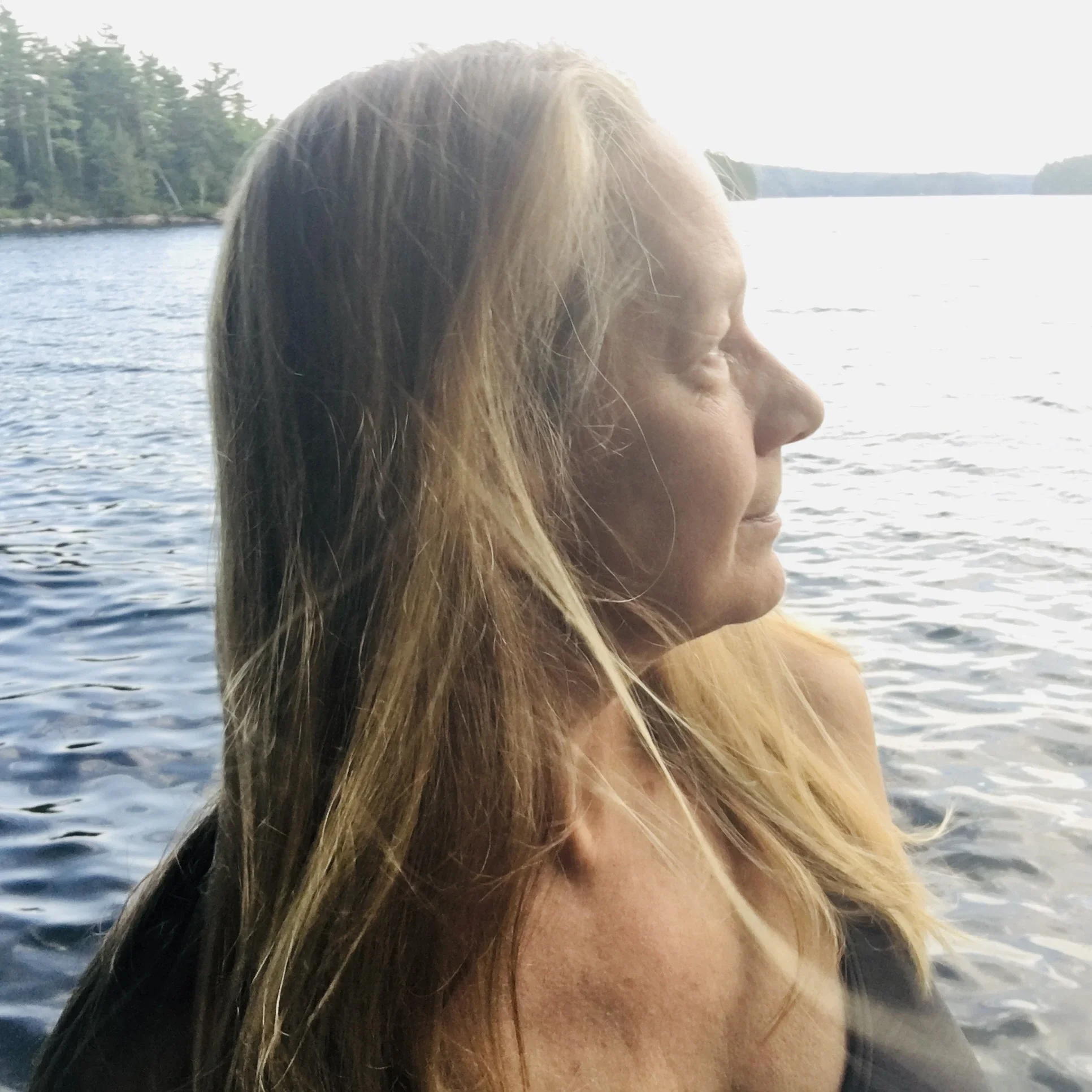 Close-up of a woman's face with long hair, appearing to be in profile, near a body of water with trees in the background, during daytime.