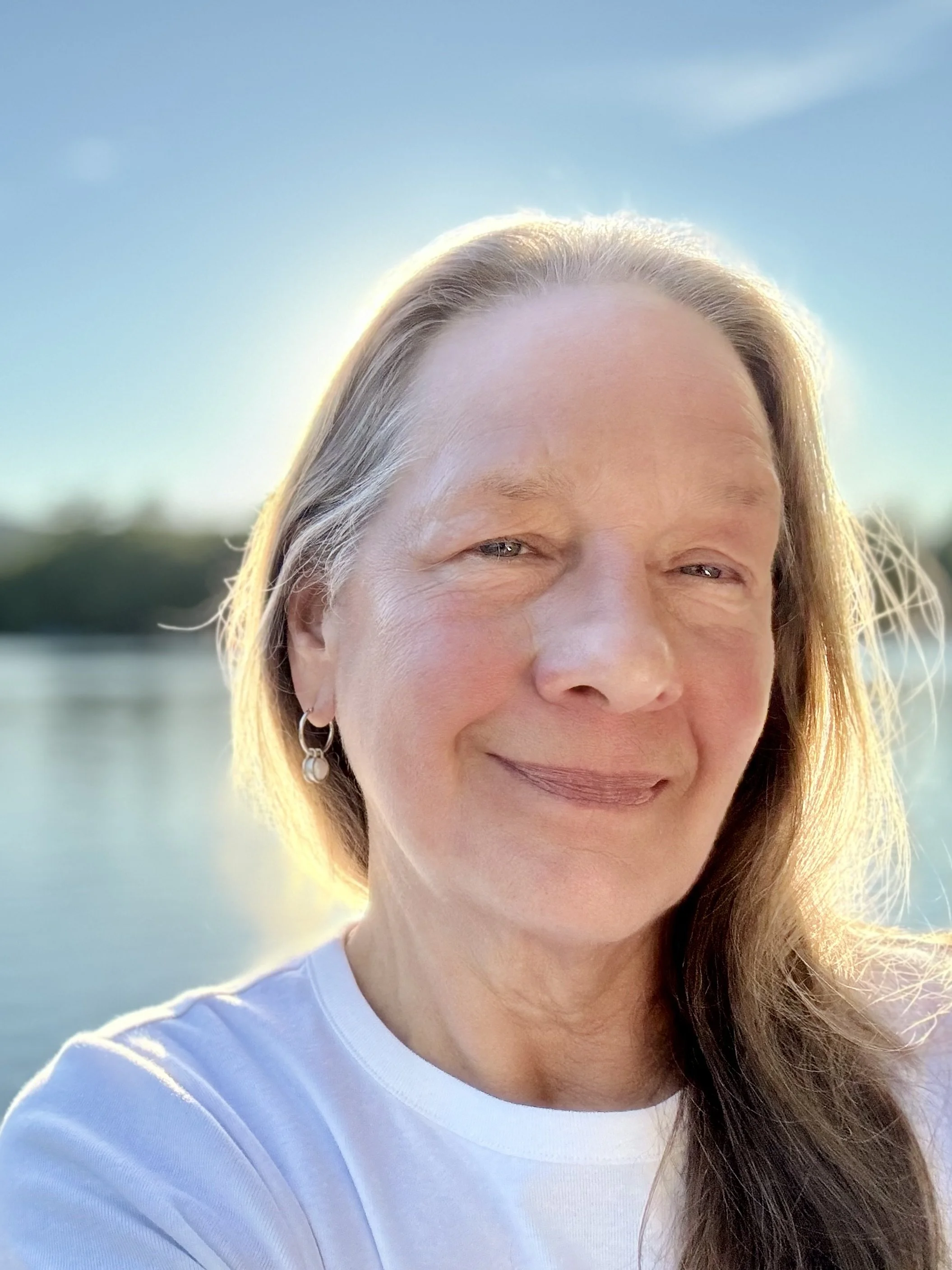 A woman with blonde hair and earrings smiling outdoors near a body of water with a clear blue sky.