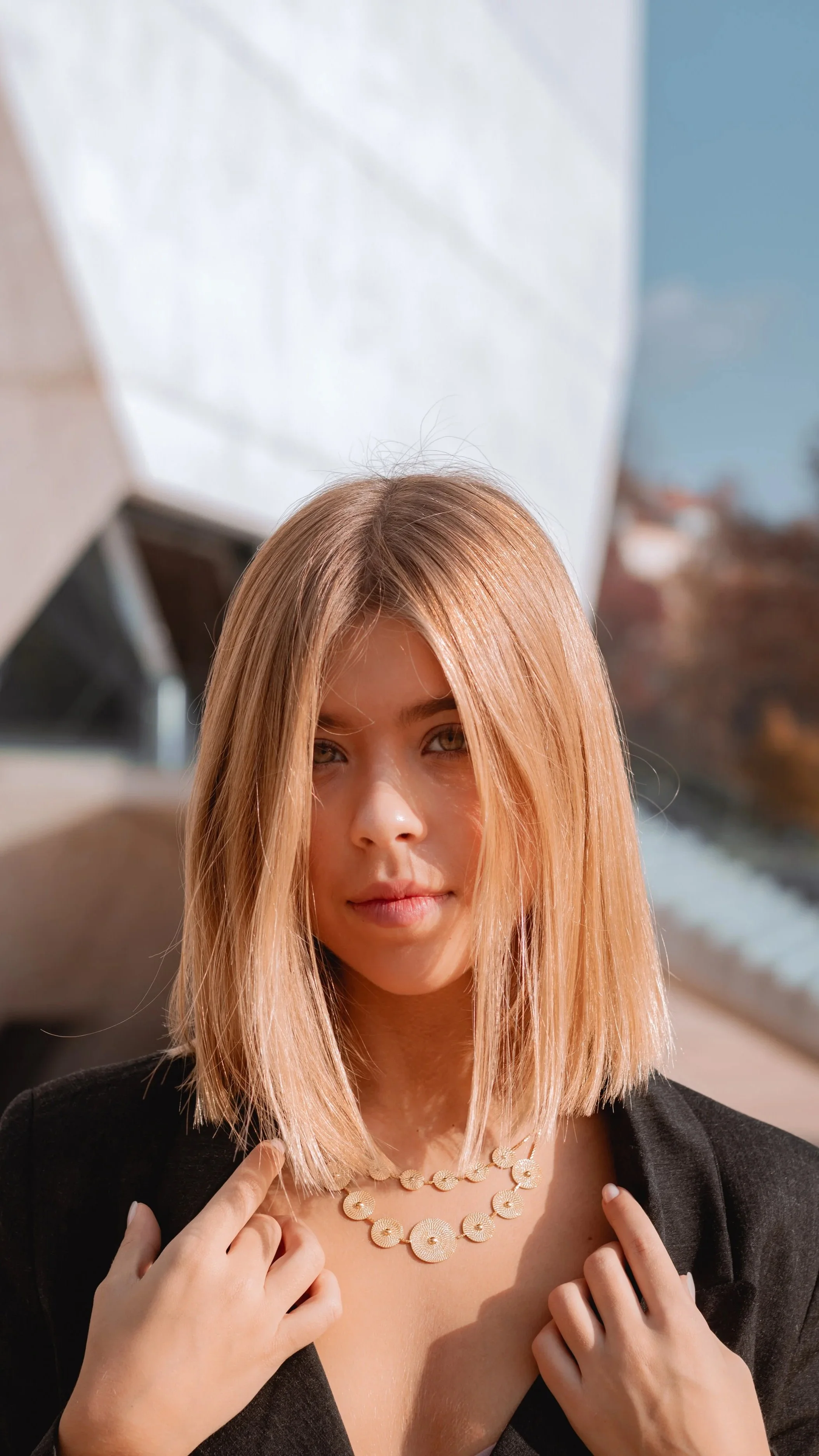 Mulher com cabelo liso, cabelo castanho claro, usando uma jaqueta preta e colar de pérolas, sorrindo levemente em ambiente externo.