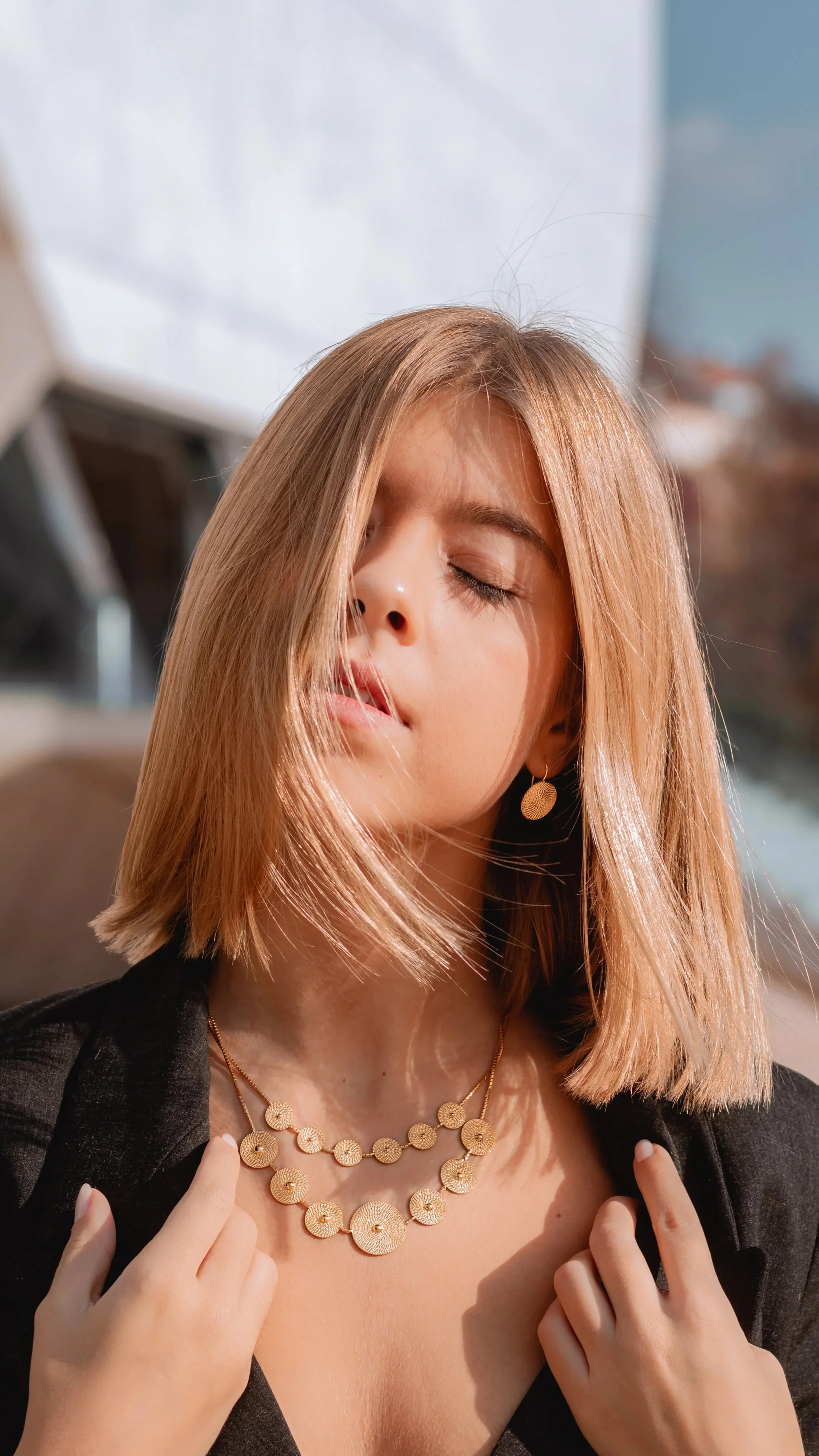Mulher com cabelo castanho claro, usando joias douradas e roupa preta, em exterior. Ela tem os olhos fechados e expressão serena.