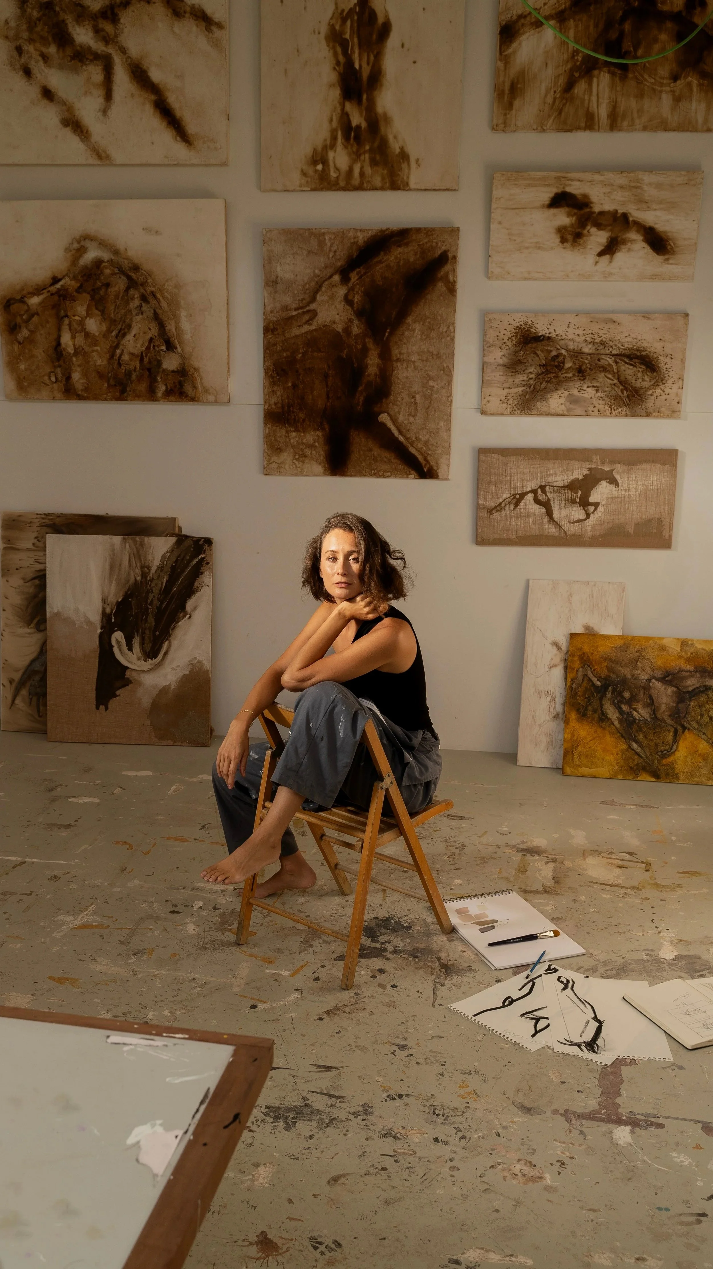 Mulher sentada numa cadeira de madeira em um estúdio de arte, rodeada por telas com pinturas abstratas em tons de marrom e bege, com materiais de pintura no chão.