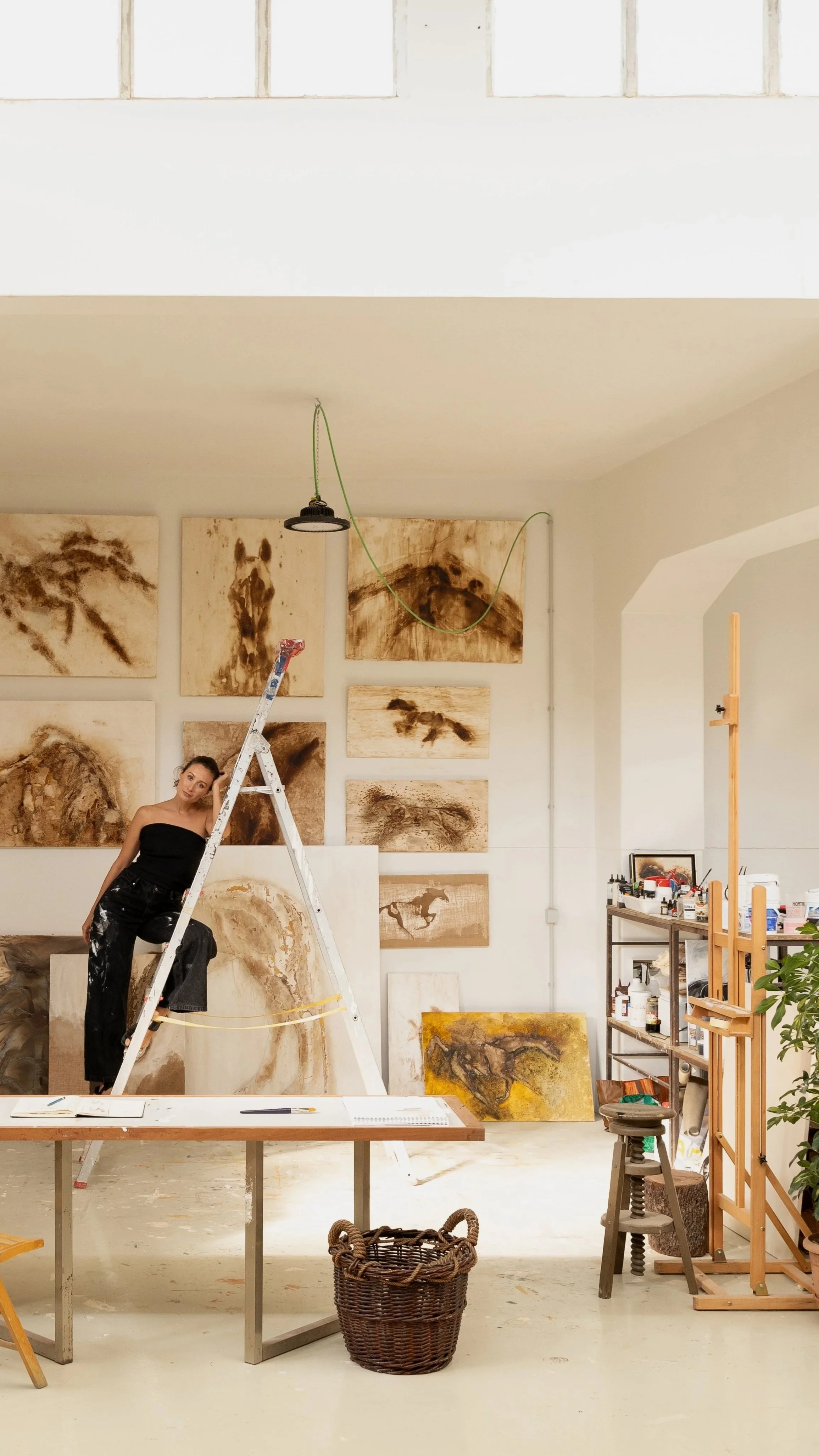 Artista em um estúdio de arte com telas de pintura ao fundo, numa escada, rodeada de materiais de pintura e uma mesa.
