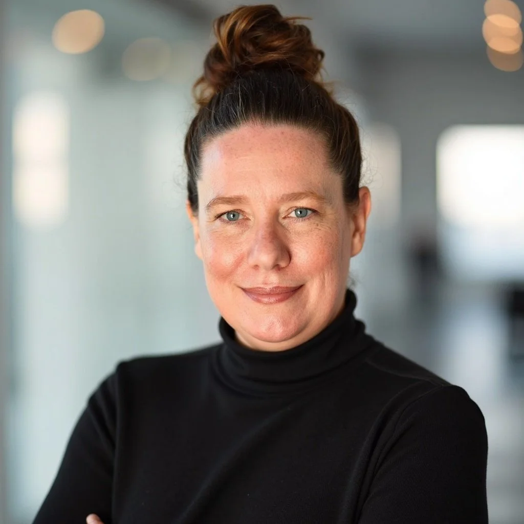 Professional woman with auburn hair in a bun, wearing a black turtleneck, smiling in an indoor setting with blurred background lighting.