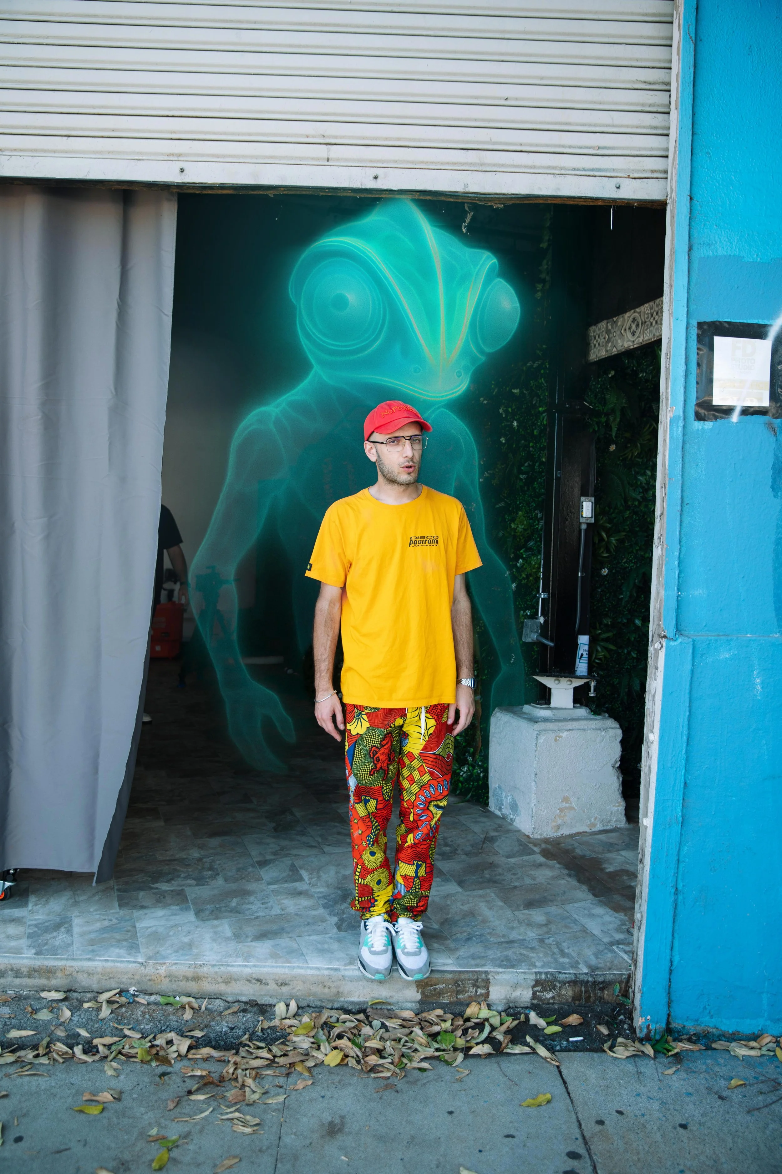 A man standing in front of a digital art display of an alien-like creature with glowing blue outlines inside a doorway with a partially open gray curtain, outdoors with leaves on the ground and a blue wall on the right.