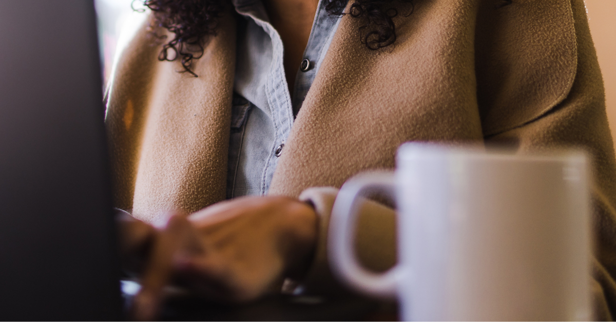 A realtor wearing a tan coat and denim shirt works on a laptop. A white coffee mug is in the foreground, creating a cozy, focused atmosphere.