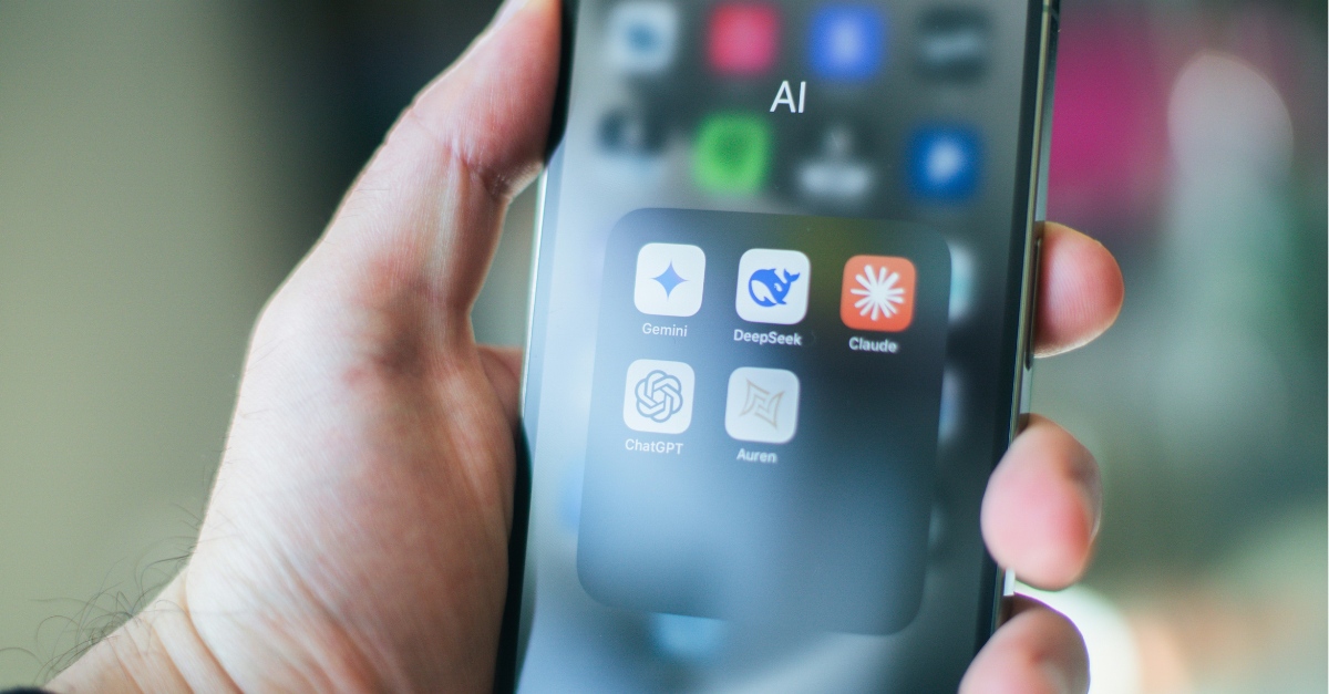 A hand holds a smartphone displaying a folder named "AI" with AI marketing tools: Gemini, DeepSeek, Claude, ChatGPT, and Auren. The background is blurred.